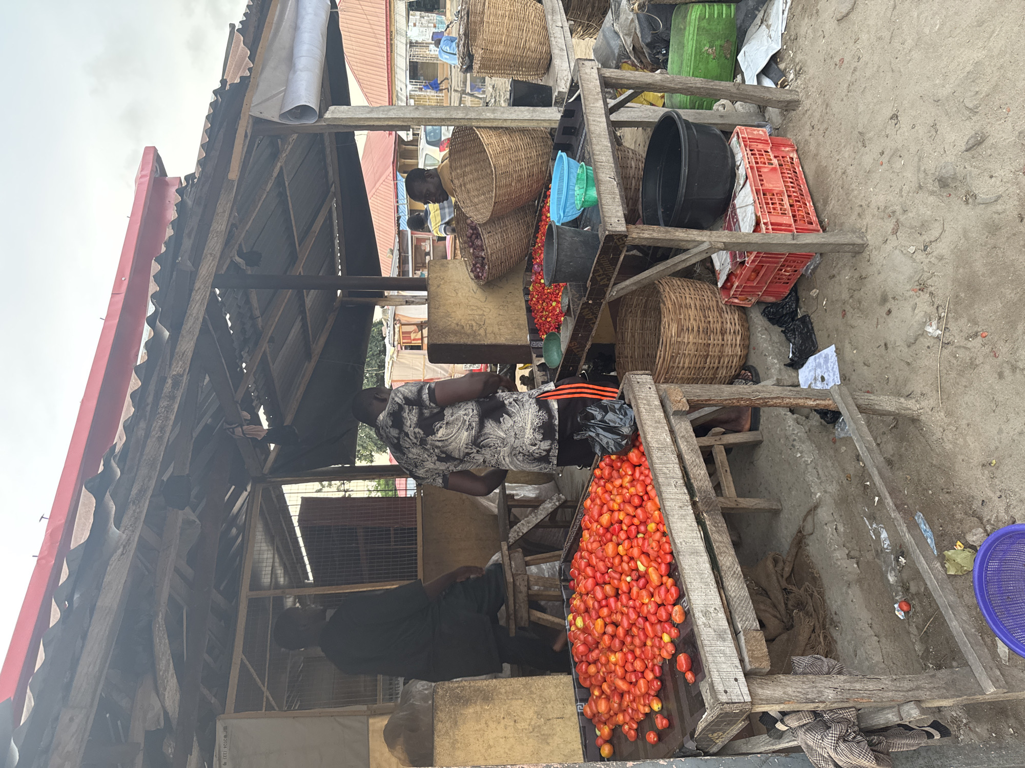 The image shows a market stall in Badagry, Nigeria. The stall has a corrugated metal roof supported by a wooden frame. A man in a patterned shirt and dark shorts is standing behind a wooden counter laden with tomatoes. Baskets of onions and other produce are displayed on a higher level behind him. Black and red crates are visible on the upper shelves. The ground around the stall is dirt and concrete.