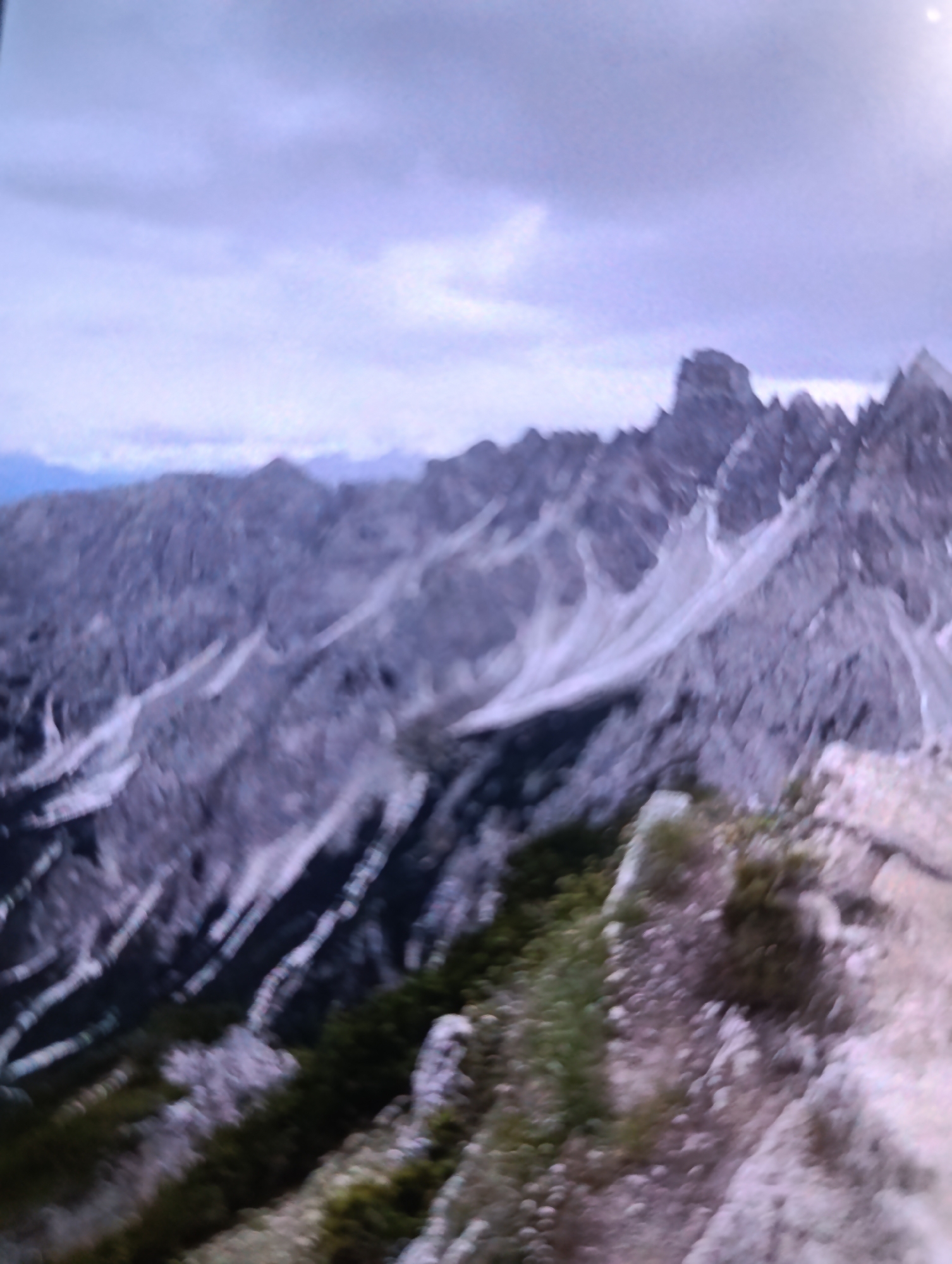 The image shows a blurred, wide-angle view of a mountainous landscape under a cloudy sky. Jagged, rocky peaks dominate the background, with patches of white snow or ice visible on their slopes. In the foreground, a rocky, vegetated ridge extends into the frame. The overall color palette is muted, with shades of gray, white, and dark green. The blurriness suggests motion or a lack of focus, making specific details difficult to discern. There are no people or identifiable man-made structures visible. The location is unknown.