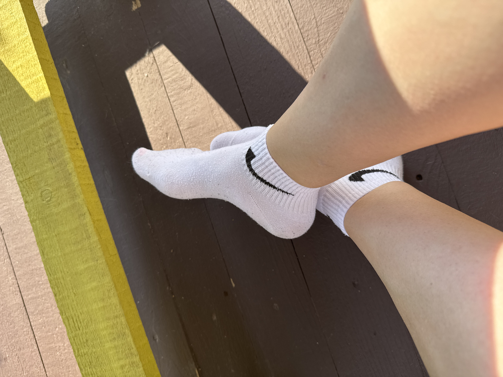 In Zav'yalovo, Russia, a close-up shot focuses on a person's legs wearing white Nike socks. The legs are positioned on a weathered wooden deck, with a yellow and green painted wooden beam running horizontally across the top of the frame. Sunlight casts distinct shadows across the wooden planks, indicating it is daytime. The scene is static, with no observable actions or interactions taking place beyond the passive presentation of the subject's legs.