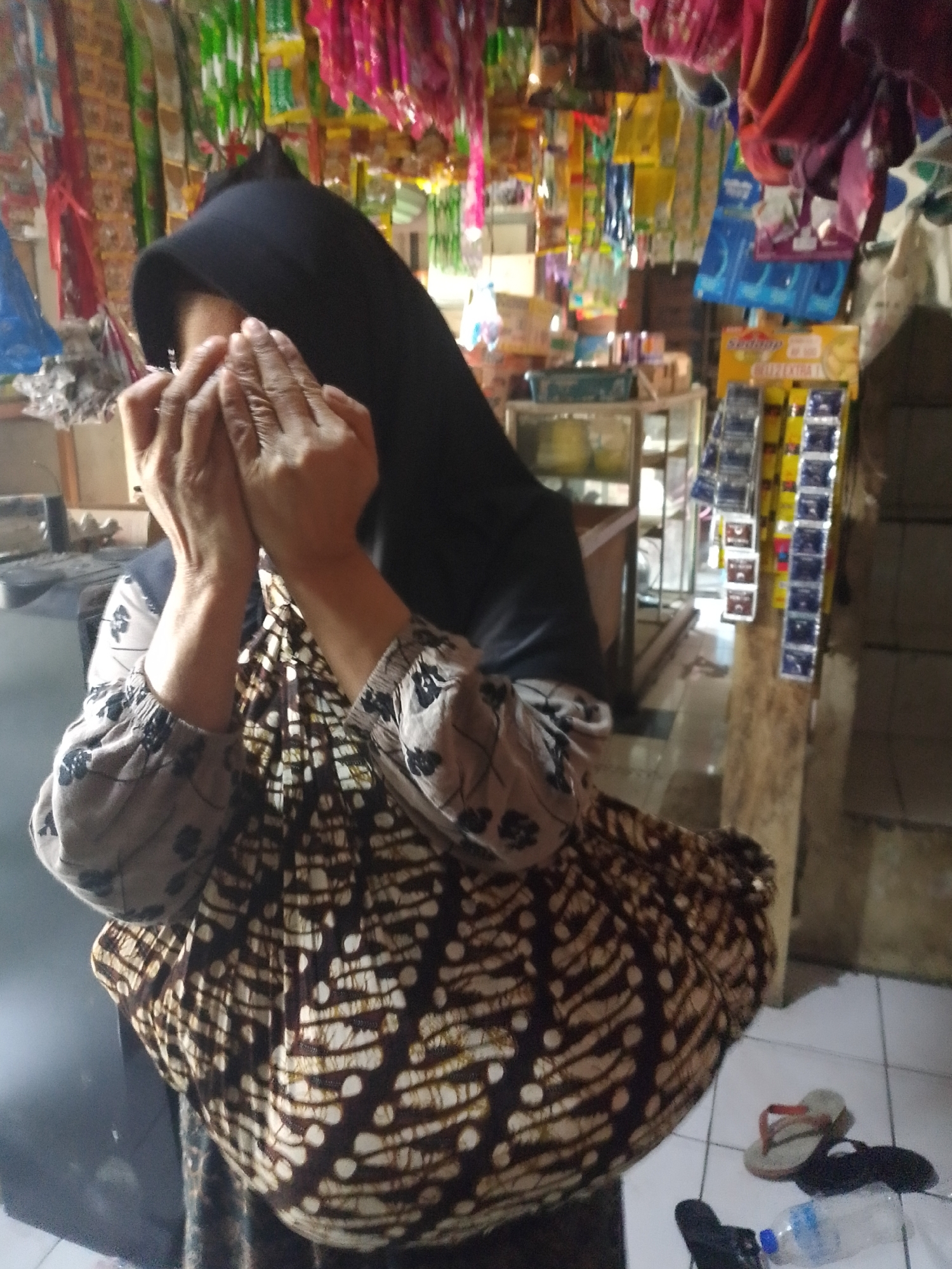 In Mukapayung, Indonesia, a person wearing a black hijab and a brown and white batik print garment is covering their face with their hands in what appears to be a small, cluttered store. The store's interior is visible, with shelves stocked with various items such as packaged goods, toiletries, and what seem to be snacks hanging from the ceiling. A glass display case is present, and a wooden post stands near the person. The floor is tiled, and on the floor, there are two pairs of flip-flops and a plastic water bottle. The lighting suggests an indoor environment, possibly with natural light from an unseen source.