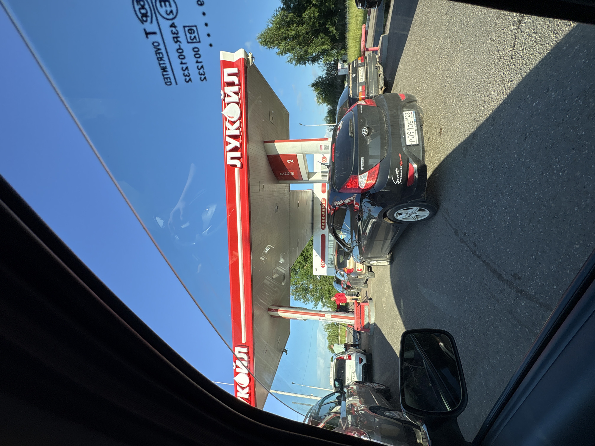 This image captures a scene at a Lukoil gas station, likely in Kirovo-Chepetsk, Russia, on a clear, sunny day. The perspective is from inside a vehicle, looking out through the side window and windshield. A line of cars is waiting to refuel, indicating a busy period at the station.
The dominant feature is the red and white canopy of the Lukoil gas station, with its prominent logo visible. Beneath the canopy are several gas pumps, with at least two cars actively refueling. In the foreground, a black Hyundai Accent is parked, facing away from the camera. Its license plate is partially visible, reading "P091OE 123". Other vehicles, including SUVs and sedans, are also in the queue.
Several individuals are present. One person, wearing a light blue shirt, is standing near a pump, possibly attending to a vehicle. Another individual, dressed in a red jacket and shorts, is seen near the pumps, appearing to be involved in the refueling process. The surrounding environment includes asphalt pavement, green trees in the background, and a clear blue sky overhead. Reflections are visible on the car window and the gas station canopy, suggesting a bright day. The overall impression is one of a typical, albeit busy, day at a Russian gas station.