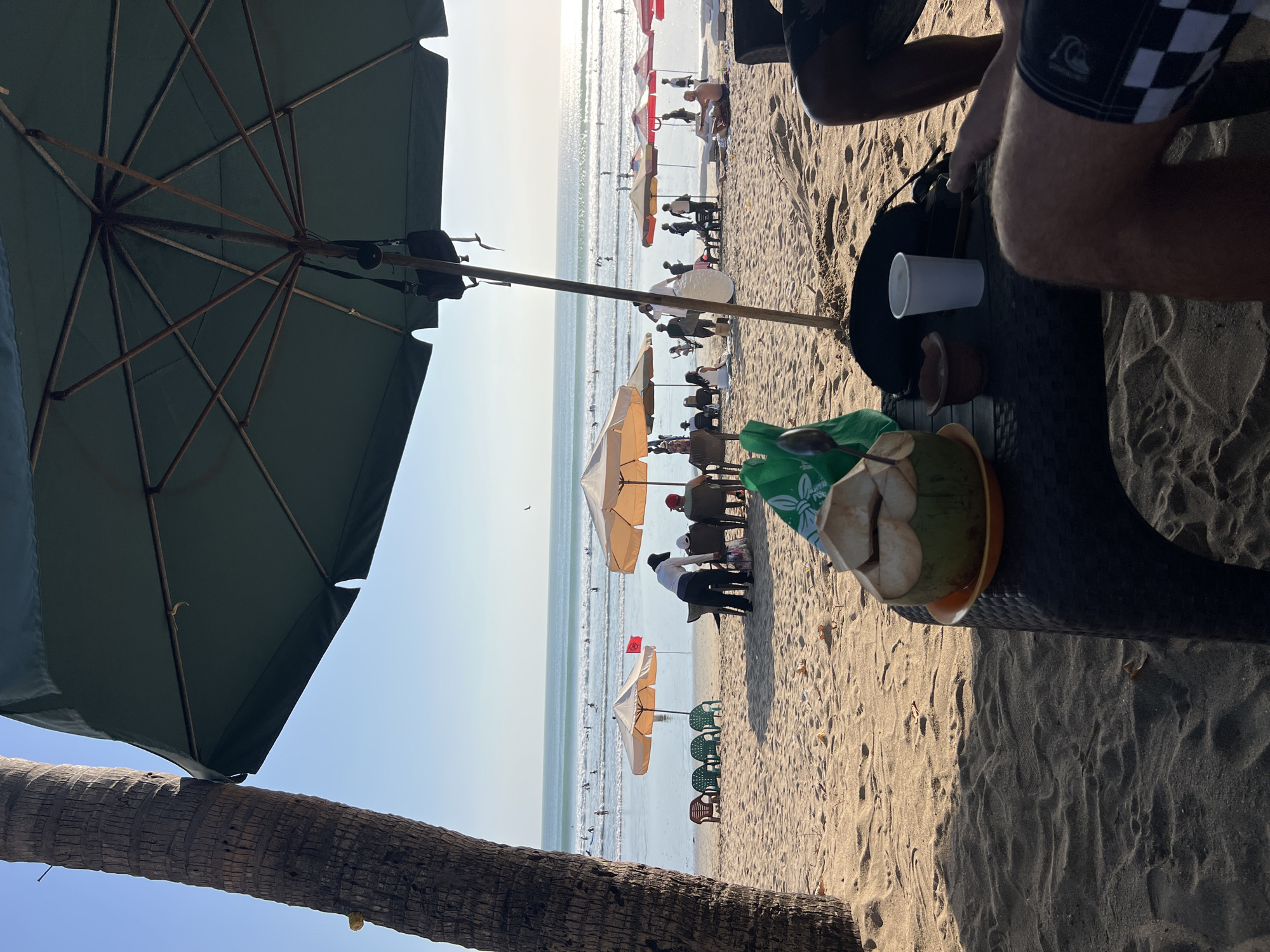 In Kuta, Indonesia, this image captures a beach scene with a focus on a table in the foreground. A half-opened coconut with a straw rests on an orange plate on the dark-colored table. To its left, a green plastic bag is visible, and a small clay pot sits next to a white paper cup. Beyond the table, a sandy beach stretches towards the ocean, dotted with several beach umbrellas and chairs. People are scattered across the beach, some sitting under umbrellas, while others are in the water, apparently surfing. The ocean appears calm with small waves, and the sky is clear and bright, indicating a sunny day. A palm tree trunk is prominent on the left side of the frame, and the shade of a large green umbrella partially covers the foreground.