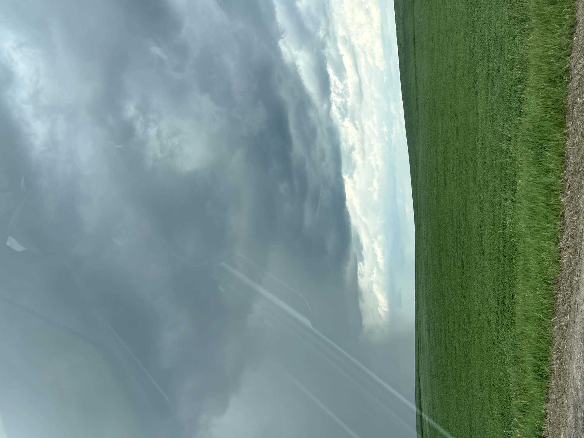 A photograph captures a scene of a large green field under a turbulent sky, likely taken from within a vehicle. The sky is filled with dark, ominous clouds, with a visible area of potential rain or storm. A portion of the photo includes what appears to be the dashboard or interior of a vehicle, suggesting the image was taken from within it. The edge of a harvested or bare ground area borders the green field. Without location specific information, I cannot determine the precise location in Голацкі сельскі Савет, Belarus.