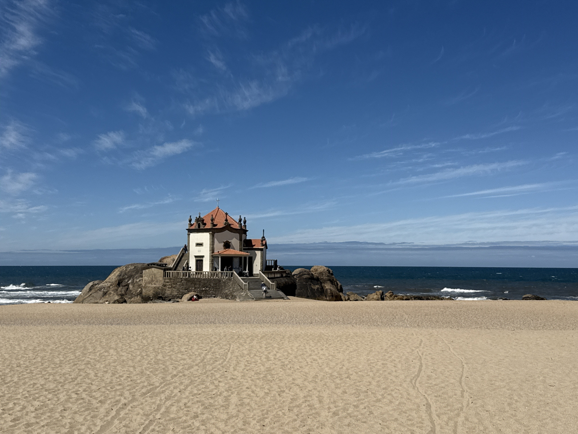 The image depicts the Capela do Senhor da Pedra, a chapel in Vila Nova de Gaia, Portugal, situated on a sandy beach. The chapel, with a red-tiled roof and white walls, sits atop a rocky outcrop, accessible by a set of stairs. The Atlantic Ocean is visible behind the chapel, with small waves breaking on the shore. The sky is blue with scattered cirrus clouds. The sandy beach stretches out in the foreground. There are no people visibly interacting in the main scene, though figures can be faintly discerned in the chapel and nearby.
