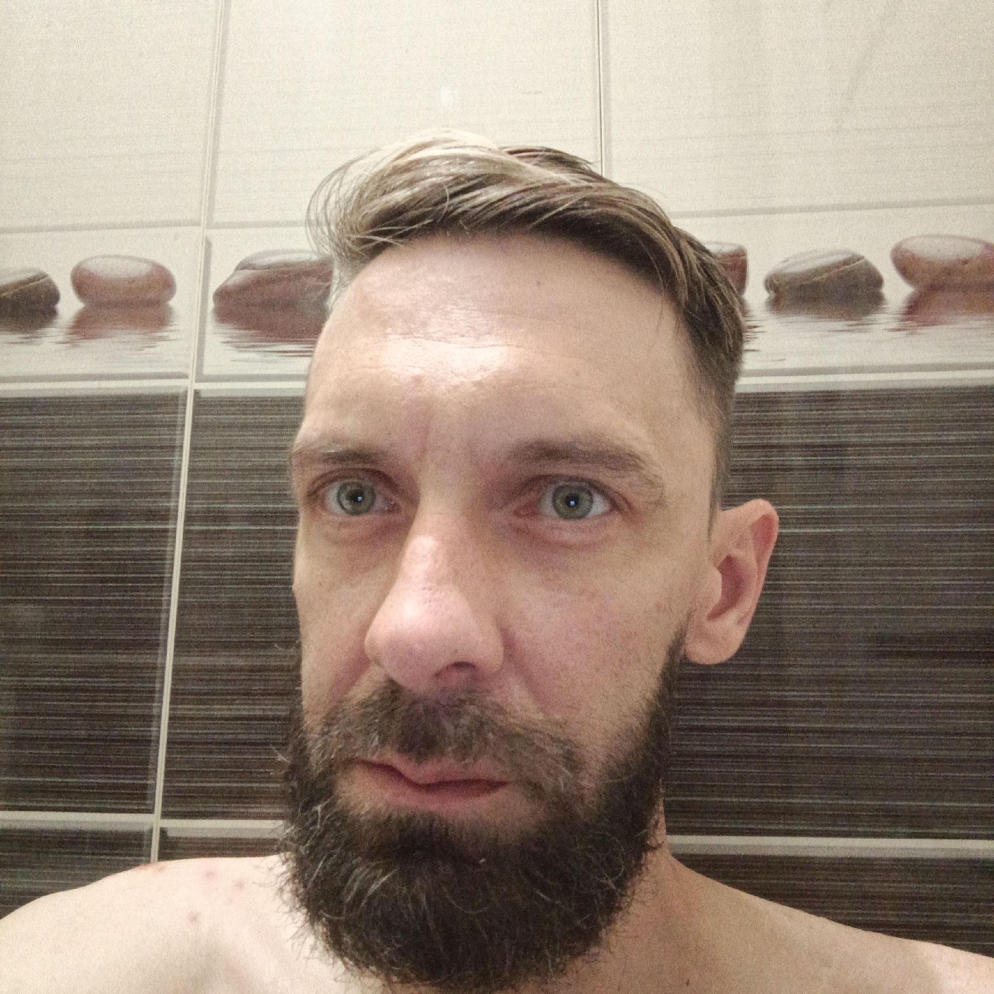A close-up, front-facing image depicts a man with light skin, blue eyes, and short, light brown hair with lighter highlights. He has a full, dark brown beard with some graying hairs. The background is a bathroom with horizontally striped brown and white tiles, and a decorative row of tiles with images of pebbles reflected in water. The man appears shirtless, and there are minor skin imperfections visible on his shoulder. The location in the city of Марковский, Russia is not discernible from the image.