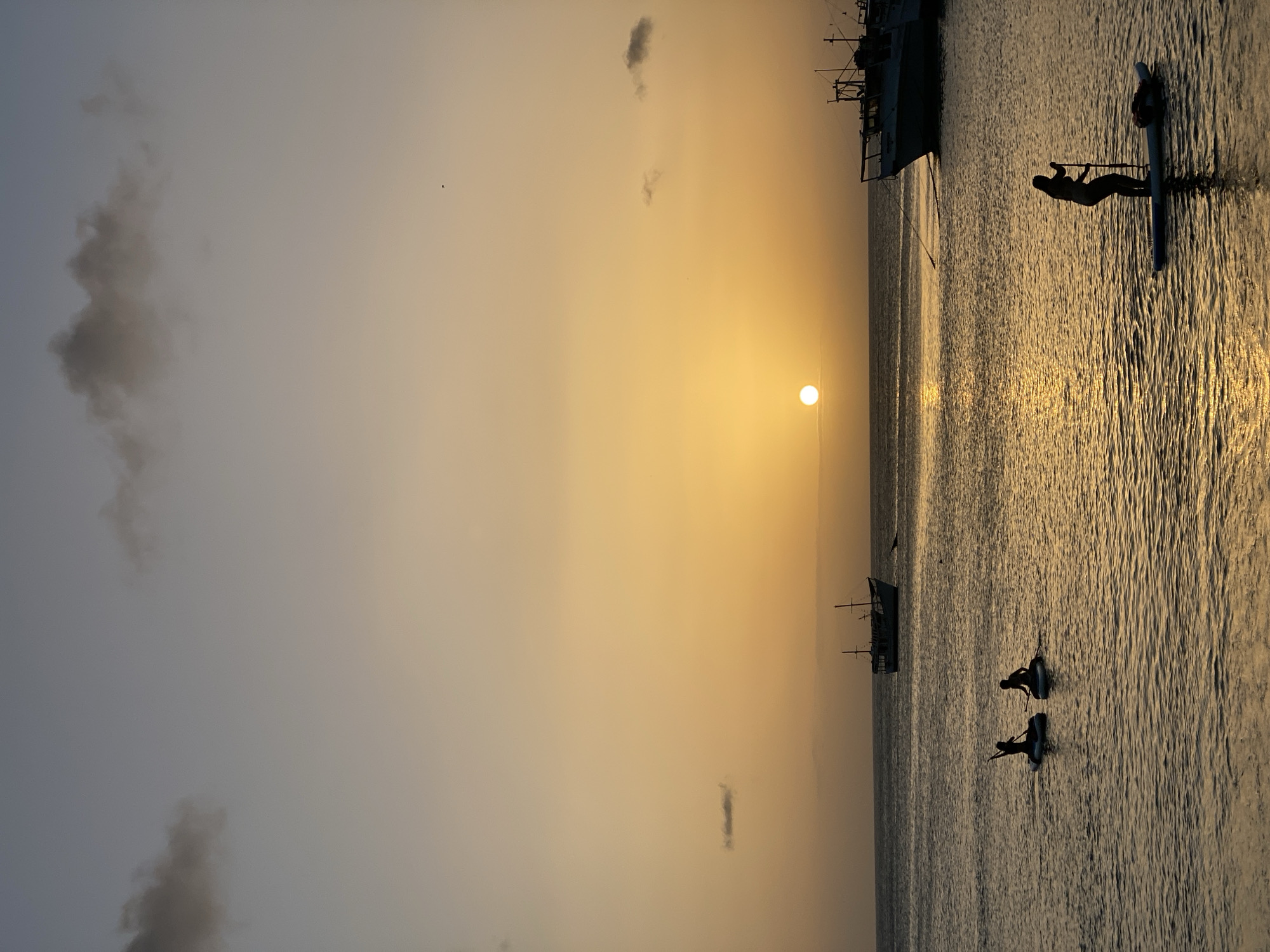 The image captures a sunset over the water in Juan Griego, Venezuela. Silhouetted figures on paddleboards navigate the calm sea. In the background, two boats are anchored, one appearing to be a larger vessel and the other a more traditional sailing ship. The sky is a gradient of orange and yellow, with a few wispy clouds present. The sun, a bright circle, is setting on the horizon, casting a golden reflection on the water.