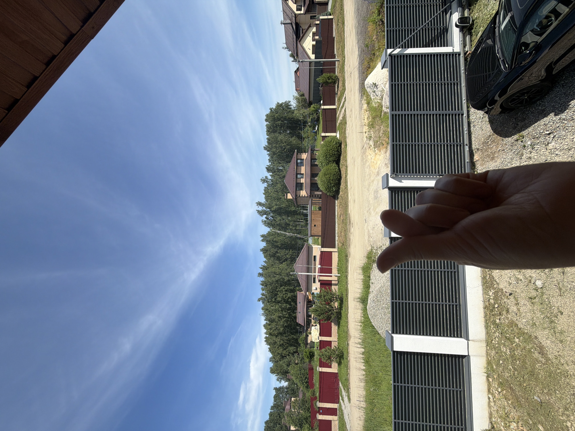 In Malinovka, Russia, a person's hand forms a finger heart gesture in the foreground, obscuring a portion of a modern, dark-colored car parked on a gravel driveway. Behind the car, a gray metal fence with white pillars runs horizontally across the midground, separating the property from a patch of overgrown grass and gravel. Beyond the fence, a row of residential houses with brown roofs and brick facades is visible, backed by a dense forest of green trees under a clear blue sky with wispy white clouds. The scene appears to be a residential area on a sunny day.