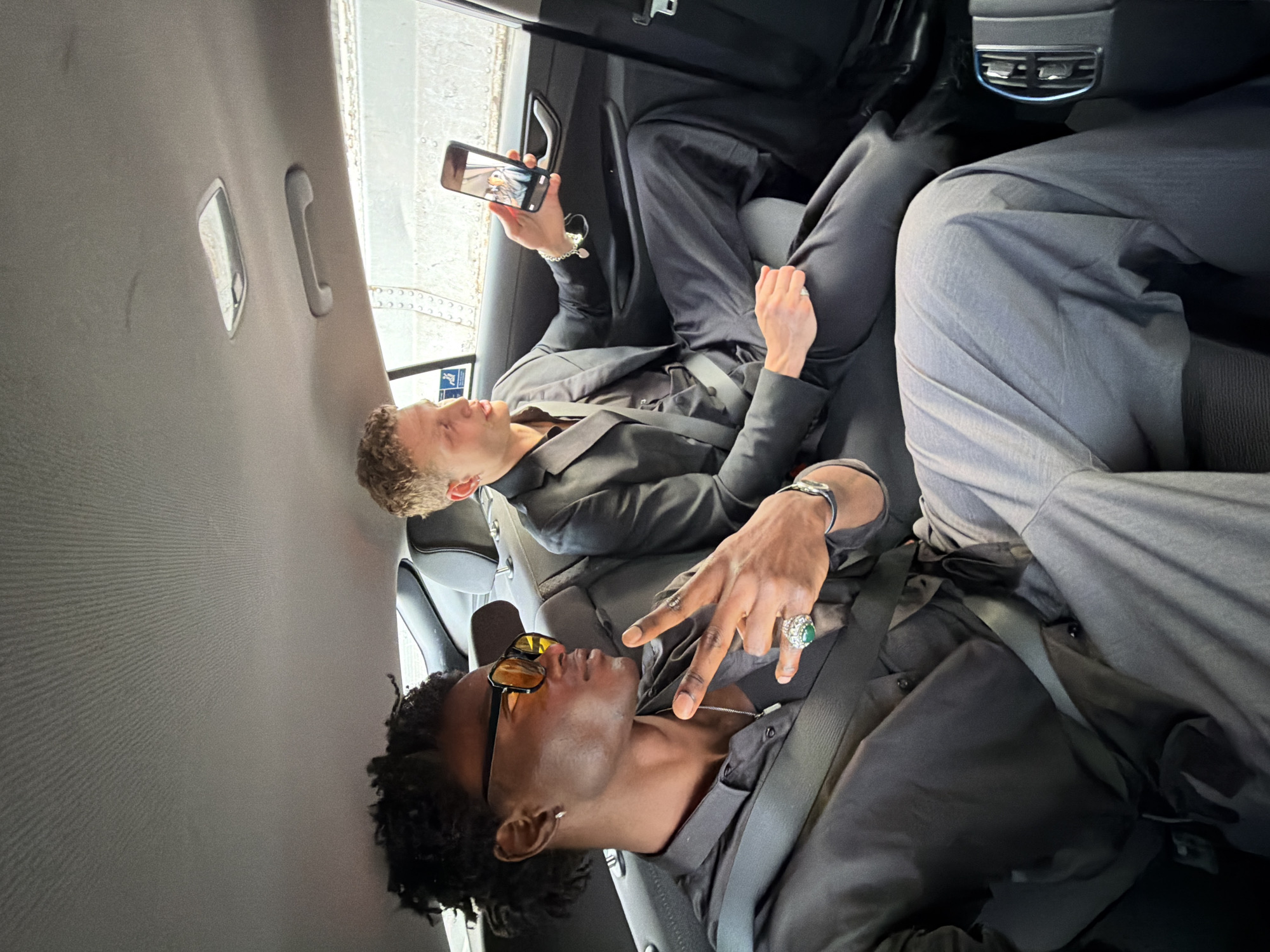 Two men are seated in the back of a vehicle during daylight. The man positioned on the left, closer to the viewer, has dark, curly hair and wears amber-tinted sunglasses, a black button-down shirt over a black undershirt, and light grey wide-leg trousers. He wears a silver hoop earring in his left ear, a silver chain with a white pendant, and a large ring with a green central stone surrounded by smaller clear stones on his right ring finger, along with a silver bracelet on his right wrist. He is making a "peace" sign with his right hand and is secured by a black seatbelt across his chest.
The man on the right, further back, has short, curly, light brown hair and is dressed in a dark suit jacket, a black collared shirt, and dark trousers. A silver bracelet with a heart-shaped charm is visible on his left wrist. He holds a black smartphone horizontally in his left hand, displaying its screen which shows an image of two faces, towards the man on the left. He is also secured by a seatbelt.
The car's interior features a grey, ribbed headliner, an overhead light fixture, and a grey grab handle above the right window. Rear air vents with metallic accents are visible on the back of the front center console. Through the rear right window, a large, light-colored structure with visible rivets or bolts, indicative of industrial or maritime infrastructure such as a ship's hull or bridge segment, is discernible. This exterior view is consistent with an industrial or port area within the city of London, United Kingdom.