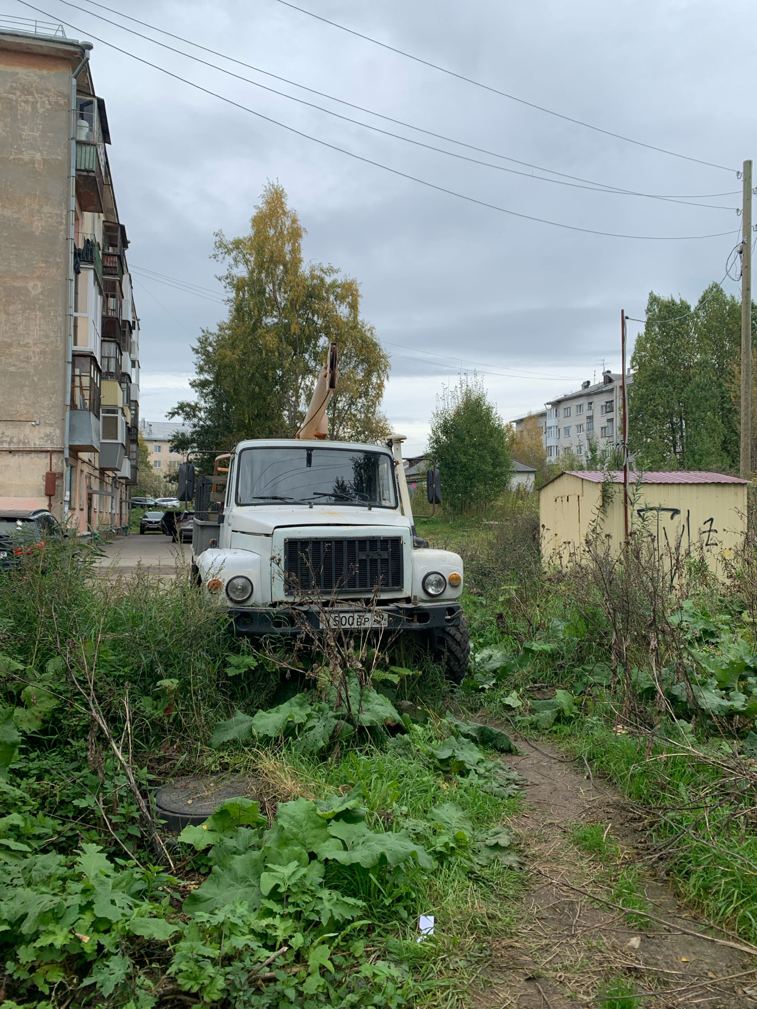 An overhead shot depicts an overcast day in Arkhangelsk, Russia, with a white truck parked on a path in front of a building. The truck, a utility vehicle with a crane, is partially obscured by overgrown vegetation. The building, a residential apartment block, is to the left of the frame, its facade weathered and peeling. A small, yellow shed stands to the right, adorned with graffiti. The ground is covered in a mix of mud and dry leaves, suggesting an autumn setting. The sky is a uniform grey, with power lines crisscrossing overhead. No people are visible in the image.