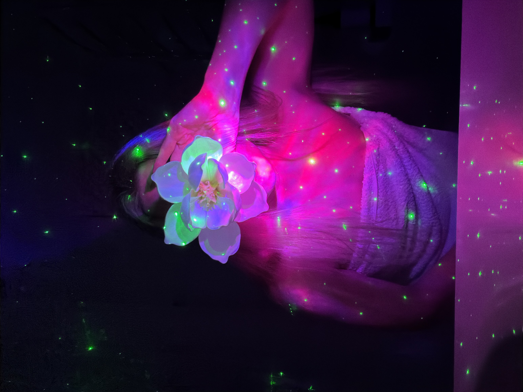 In a dark, ethereal setting in Votkinsk, Russia, a person lies reclined, primarily illuminated by vibrant, neon-like lighting. The subject's head is turned, with long hair cascading, and a hand gently holds a large, luminous flower near their face. The flower, with its petals glowing in shades of white, light purple, and green, becomes a central focal point.
The person's exposed skin on the arm, shoulder, and upper chest is bathed in vivid pink and purple light. A fluffy, light-colored towel or fabric is draped over one shoulder, also catching the colorful light. The entire scene is dotted with numerous small, bright green specks of light, resembling stars or laser projections, scattered across the person's body, the flower, and the deep black background. These green lights create a magical, cosmic, or dreamlike atmosphere.
The low-light environment suggests nighttime or an indoor space specifically designed with this type of lighting. There is no visible text in the image. The overall impression is one of serene beauty, fantasy, and artistic illumination.