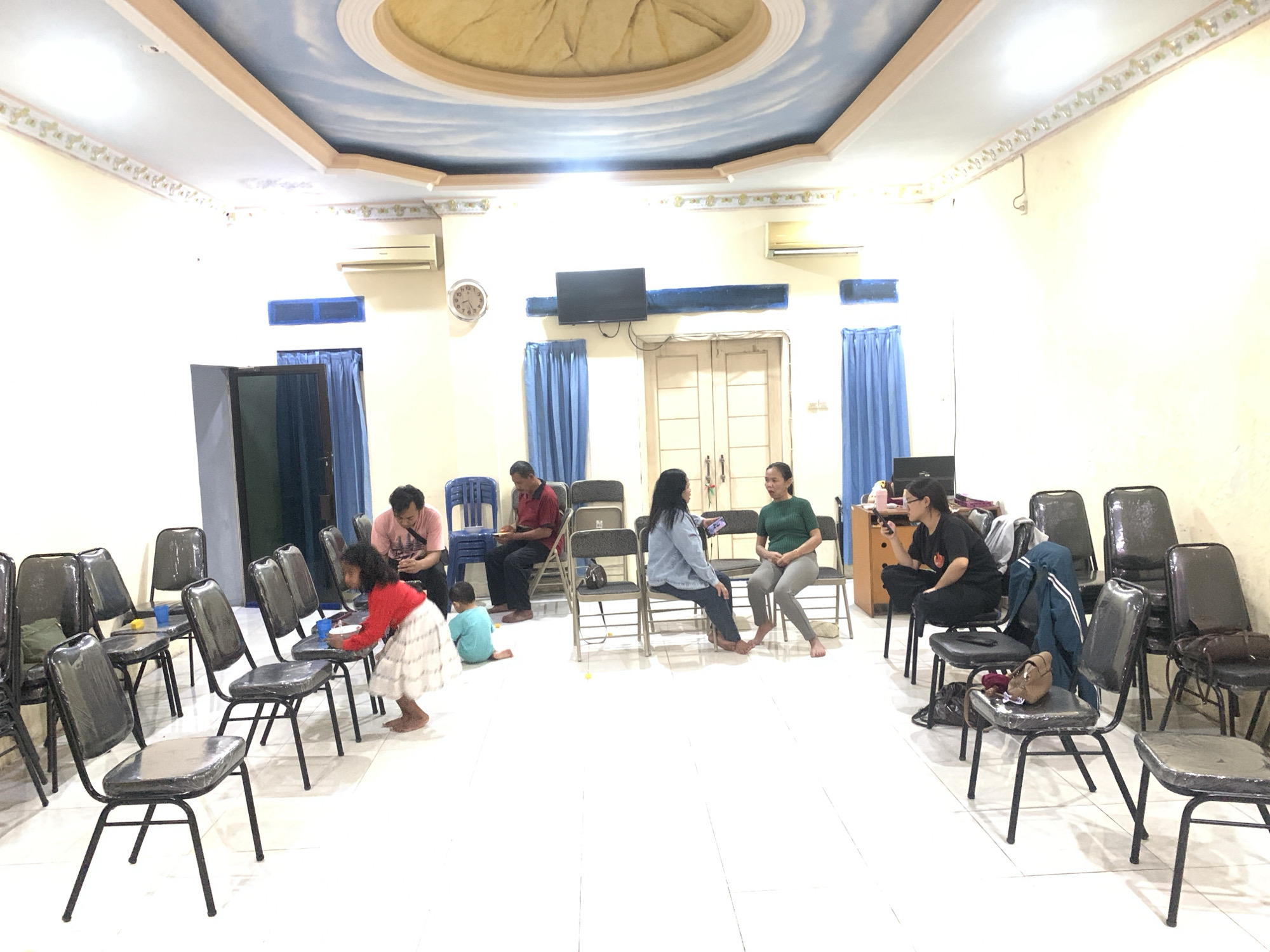 A medium shot captures an indoor gathering in what appears to be a community hall or church in Dukuh Mojo, Indonesia. The room is brightly lit, with fluorescent lights illuminating the tiled floor and cream-colored walls. The ceiling features a decorative sky mural with clouds and a central circular element. Several individuals are present, seated and standing.
In the foreground, a young girl in a white and red outfit is leaning over to pick up a white plate and a blue cup from a black plastic chair. To her right, a young child in a blue t-shirt sits on the floor, facing away from the camera.
Further back, a man in a pink t-shirt and dark pants is seated, looking down. Beside him, a man in a red polo shirt and dark pants is also seated. Two women are engaged in conversation. One woman, wearing a blue denim jacket and dark pants, sits on a black chair, holding a phone. The other woman, in a green t-shirt and grey pants, is seated on a black chair, facing the woman in denim. Another woman in a black t-shirt and dark pants is seated on a black chair, looking at her phone. Several black plastic chairs are arranged throughout the space, some empty and some occupied. Blue curtains are visible behind some of the individuals.
On the back wall, two air conditioning units are mounted, and a black television is fixed to the wall above a blue bar. A clock with a circular face and Roman numerals is visible on the wall. A wooden cabinet or desk is partially visible on the right side of the image. Bags are placed on some of the chairs. The overall atmosphere suggests a casual meeting or event.