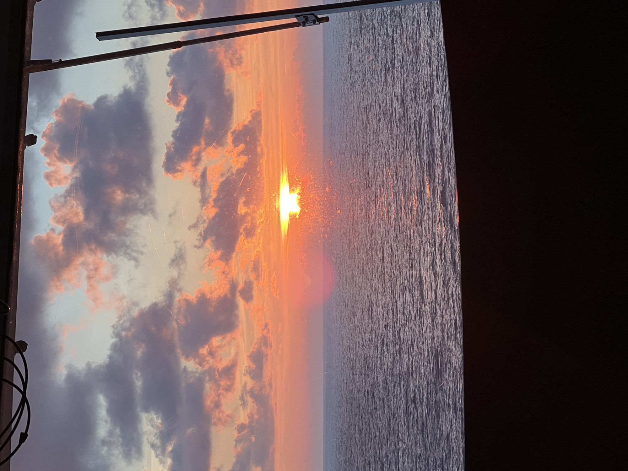 A sunset over the ocean is depicted. The sun is low on the horizon, casting a bright orange glow that is reflected on the water. The sky is filled with colorful clouds, ranging from pink and orange to purple and gray. The ocean is calm, with gentle waves. The image appears to be taken from a boat or a window, as there is a dark foreground and what looks like a metal railing on the right side of the frame. There are no people or animals visible in the image. The location is Unknown, Unknown.