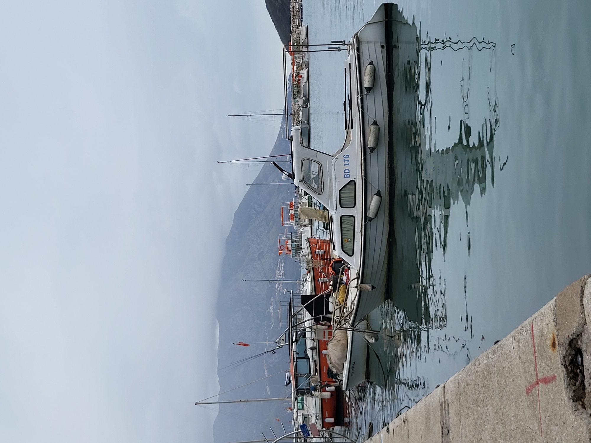 The image depicts a harbor scene in Budva, Montenegro, under an overcast sky. The foreground features a concrete pier edge on the left, exhibiting some red markings and a damaged section. The water is calm, reflecting the moored vessels.
The central subject is a light grey motorboat, registered "BD 176," featuring a small cabin and several white fenders along its port side. To its immediate left, another smaller light-colored boat, bearing the registration "777BD," is partially visible with a large, light-colored sack covering its bow area. Further left and in the background, a larger orange-hulled motorboat with a white cabin and mast is visible, displaying an orange flag and several fishing rods mounted on its stern.
In the mid-ground and background, numerous other boats, including sailing yachts with tall masts and smaller motorboats, are moored across the harbor. A mountainous landscape, partially obscured by low clouds, forms the distant background, with scattered buildings visible on its slopes. A stone breakwater is discernible on the far right horizon. No individuals are present in the scene.