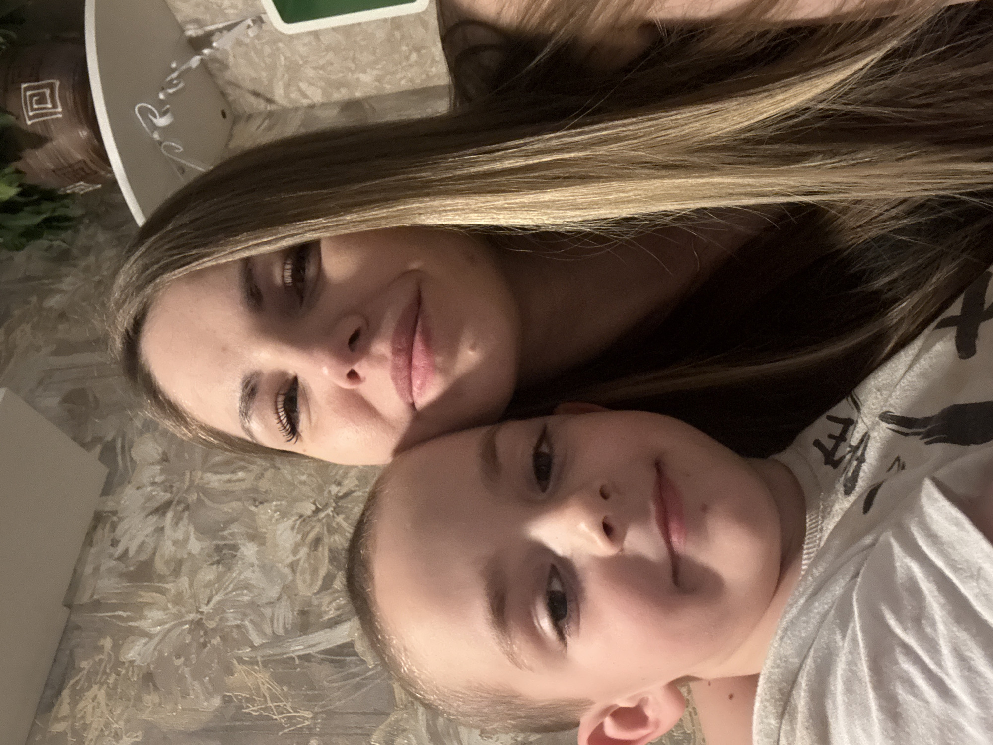 A selfie-style image shows a woman and a young boy. The woman has long, highlighted hair, fair skin, and noticeable eyelashes. The boy has short hair and is wearing a patterned shirt. The background appears to be a textured surface, possibly a bedspread or wall covering. No information in the image identifies a location in Раздолье, Russia.