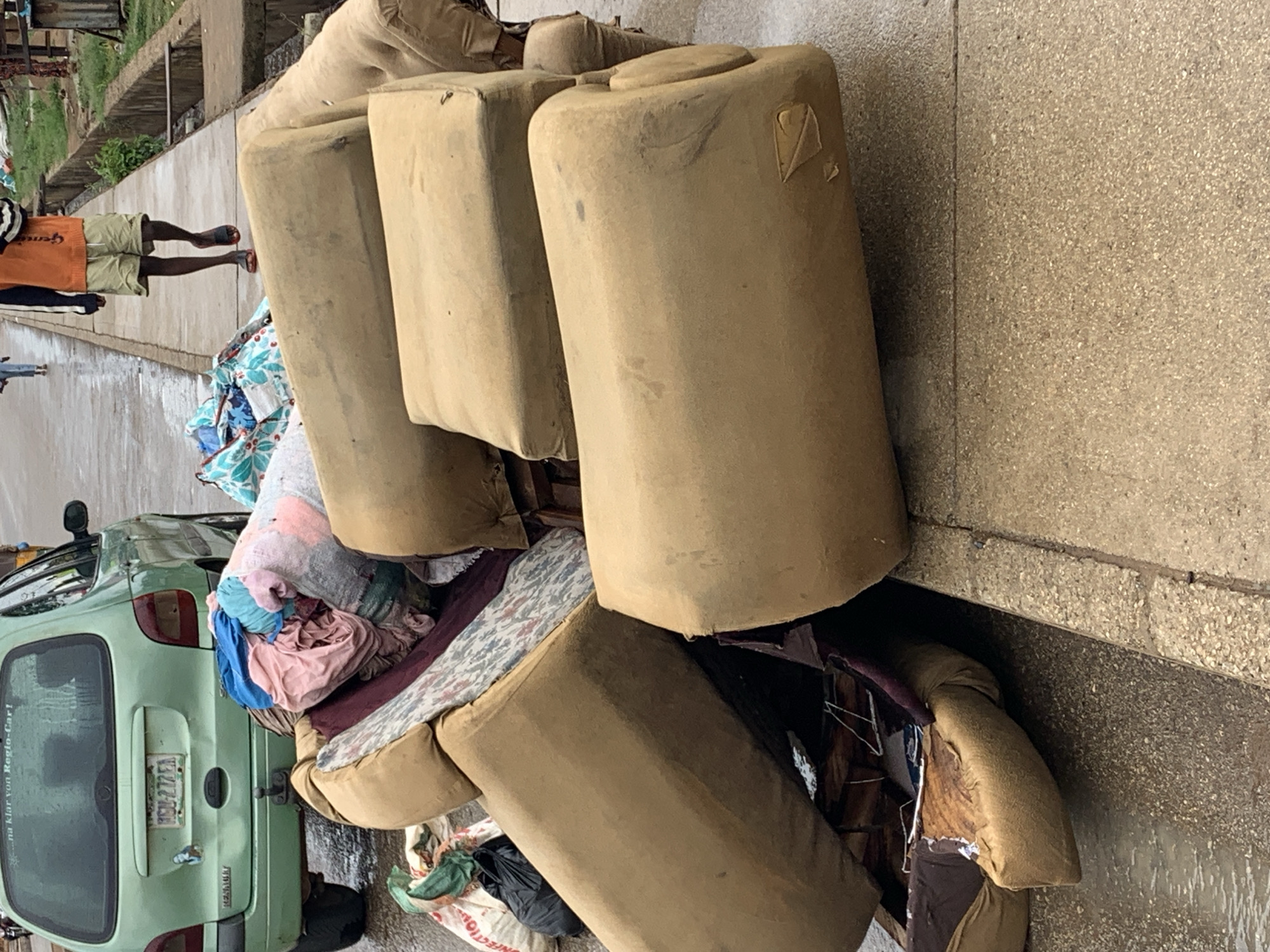 A car filled with household items, including couch cushions, is parked on the side of the road in Jos, Nigeria. A person is walking on the sidewalk. The scene appears disorganized.
