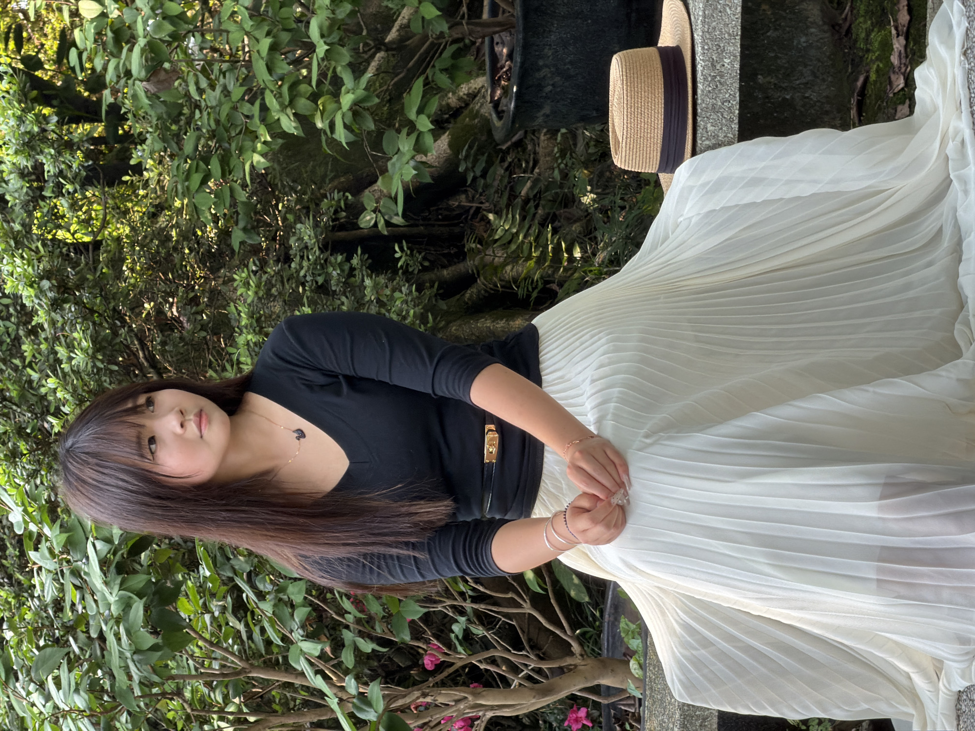 A young East Asian female is depicted seated on a grey stone bench outdoors in a garden or park setting, characteristic of Yuzhong District, China. She wears a black long-sleeved V-neck top, a thin black belt with a gold-toned buckle, and a long, pleated white skirt. Her long, straight, dark brown hair frames her face with bangs. Her expression is contemplative as she gazes towards her right (viewer's left). Her hands are clasped in her lap, displaying manicured nails and several pieces of jewelry, including a gold-toned necklace with a black swan pendant, multiple bracelets on both wrists, and rings. To her right on the bench, a straw boater hat with a dark band is placed. The background consists of abundant green foliage from various trees and shrubs, including a plant with pink blossoms on the left and visible tree roots on the right, along with dark planter pots. Natural daylight illuminates the scene.