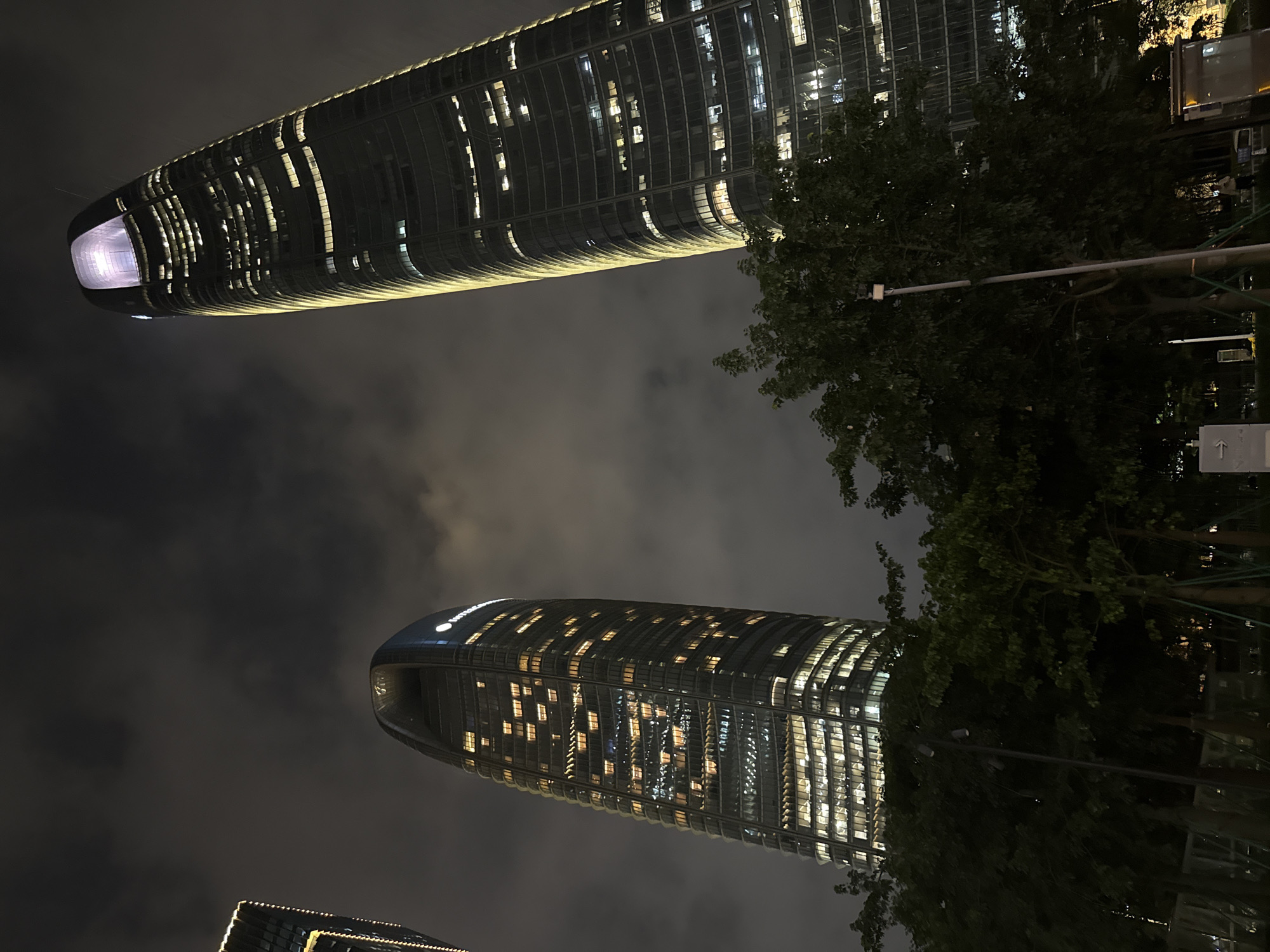 This media file showcases the nighttime cityscape of Haizhu District, China, featuring two prominent, illuminated skyscrapers. The buildings are modern and tall, with curved, glass facades reflecting the warm light emanating from their interior windows. One skyscraper, positioned higher and closer to the viewer, has a distinctive rounded top with a glowing purple light. The other, lower and further away, also displays a sleek, curved design with numerous illuminated windows.
The scene is set against a dark, cloudy night sky, creating a dramatic contrast with the lit-up architecture. Lush green trees are visible in the foreground and mid-ground, their dark silhouettes adding depth and texture to the urban landscape. Streetlights and other urban infrastructure, like a lamppost, are also present, hinting at the bustling environment. The overall mood is one of urban grandeur and nocturnal activity, with the lights of the buildings suggesting ongoing life and work within them. No specific people or activities are discernible. The time of day is clearly night. There is no visible text in the frame.