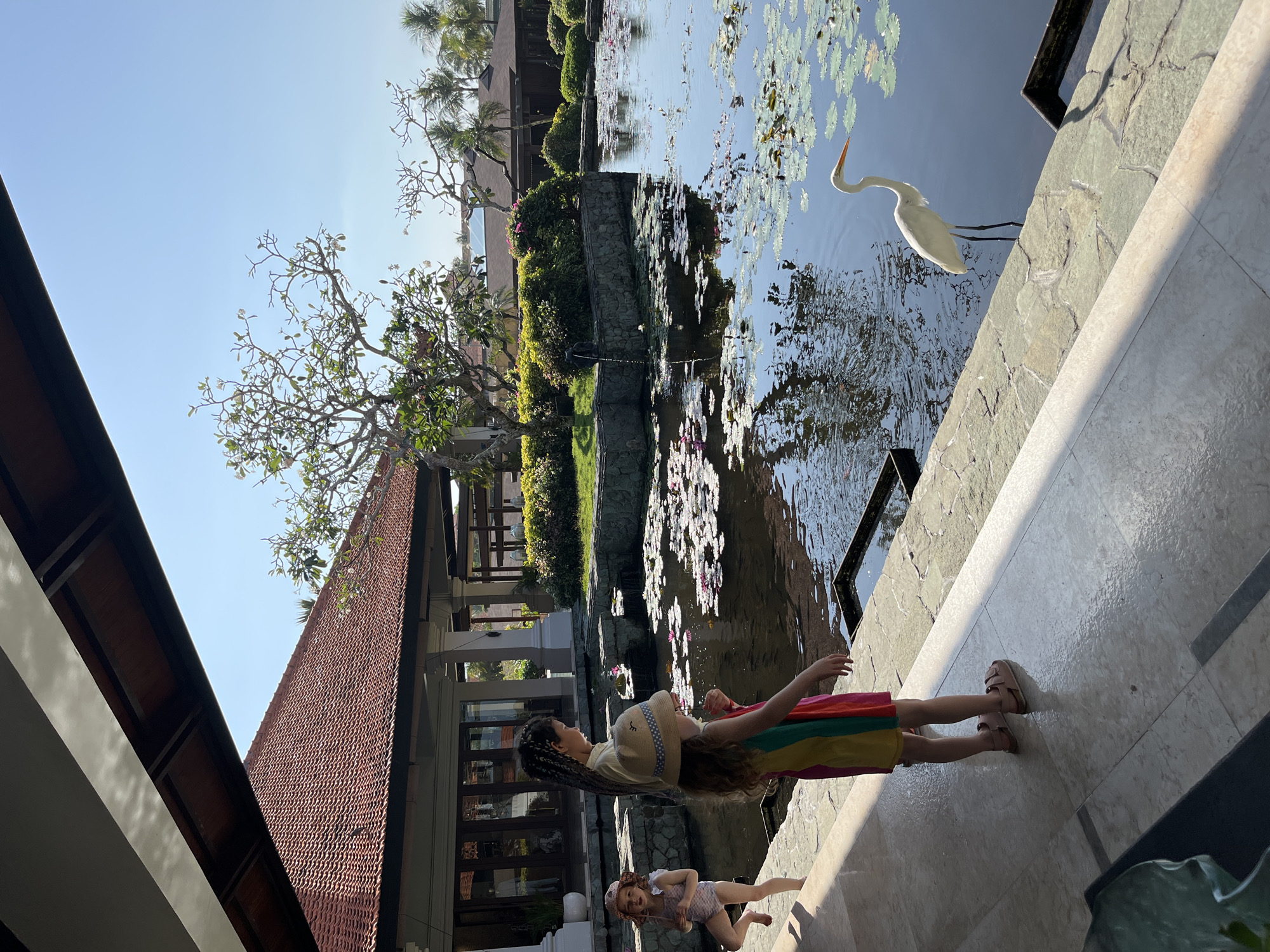 The image depicts an outdoor scene with a pond filled with water lilies and a white egret standing in the water near the edge. Three young girls are present on a marble walkway by the pond. The girl on the left is wearing a light-colored swimsuit and a sun hat. The girl in the middle is wearing a hat and a yellow top, while the girl on the right is wearing a brightly colored dress. In the background, there is a building with a red tile roof, lush greenery, and palm trees. The sky is clear and blue, suggesting a sunny day. The location appears to be a resort or a similar establishment in Kuta Selatan, Indonesia, likely a hotel with a water feature.
