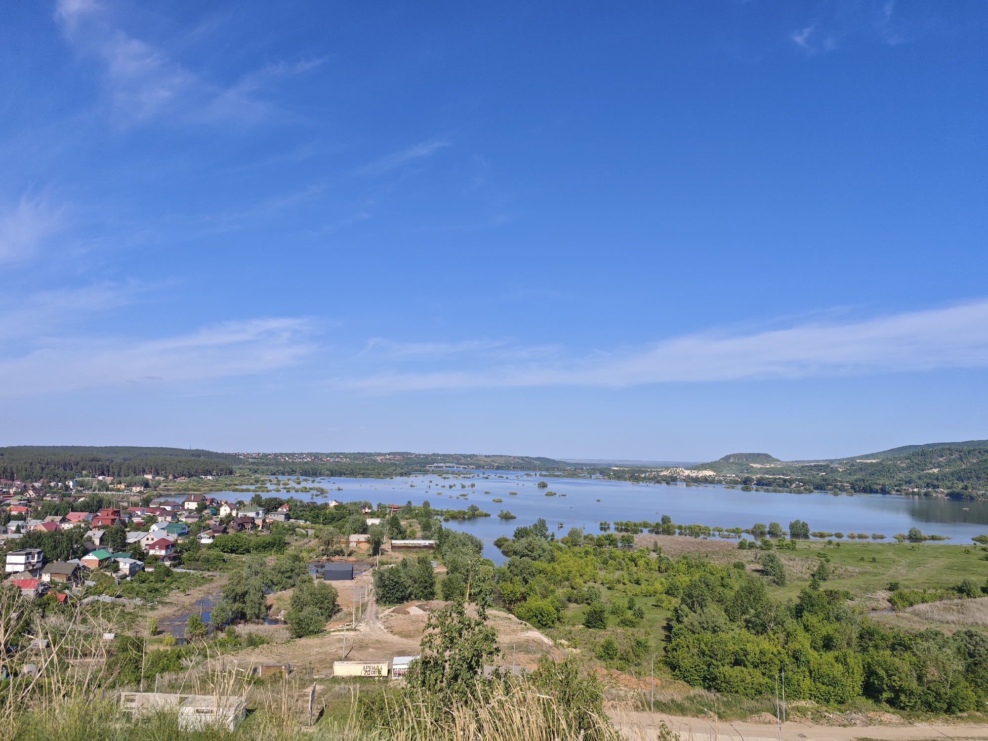 The image captures a scenic view of a landscape in Volzhskiy, Russia, encompassing a blend of urban and natural elements. The left side features a residential area with houses of various colors, mainly red and green roofs, nestled among trees. The central part of the image transitions into a body of water, likely a river or lake, dotted with numerous small islands of vegetation. The water reflects the clear blue sky, which dominates the upper half of the image, featuring a few wispy clouds. The foreground is characterized by earthy tones, with patches of dry grass and low-lying vegetation leading up to the view. The right side presents a gradual slope of land covered in greenery, transitioning into the water's edge. The overall impression is that of a tranquil, slightly elevated vantage point looking over the town and its surrounding natural environment.