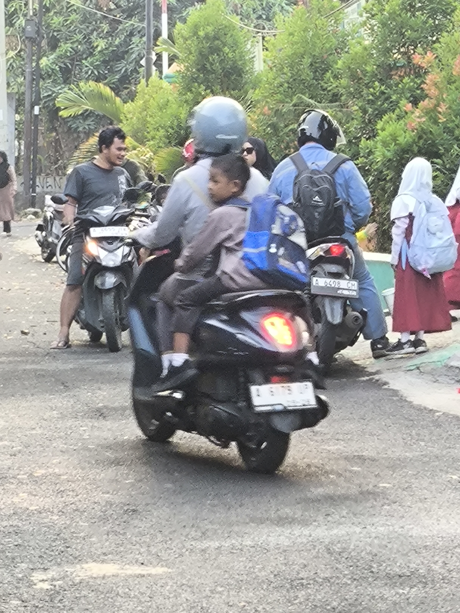 In Cilegon, Indonesia, a group of people are on motorcycles on a paved road, with trees and buildings in the background. A man wearing a dark t-shirt and shorts, with sandals, is riding a motorcycle. Behind him, another person is riding with a child. The child is wearing a gray shirt and dark pants, and is holding onto a backpack. A person in a blue shirt and black backpack is also on a motorcycle. A person wearing a hijab and a red dress, carrying a blue backpack, is standing on the side of the road. The scene appears to be during the daytime, with sunlight filtering through the trees.