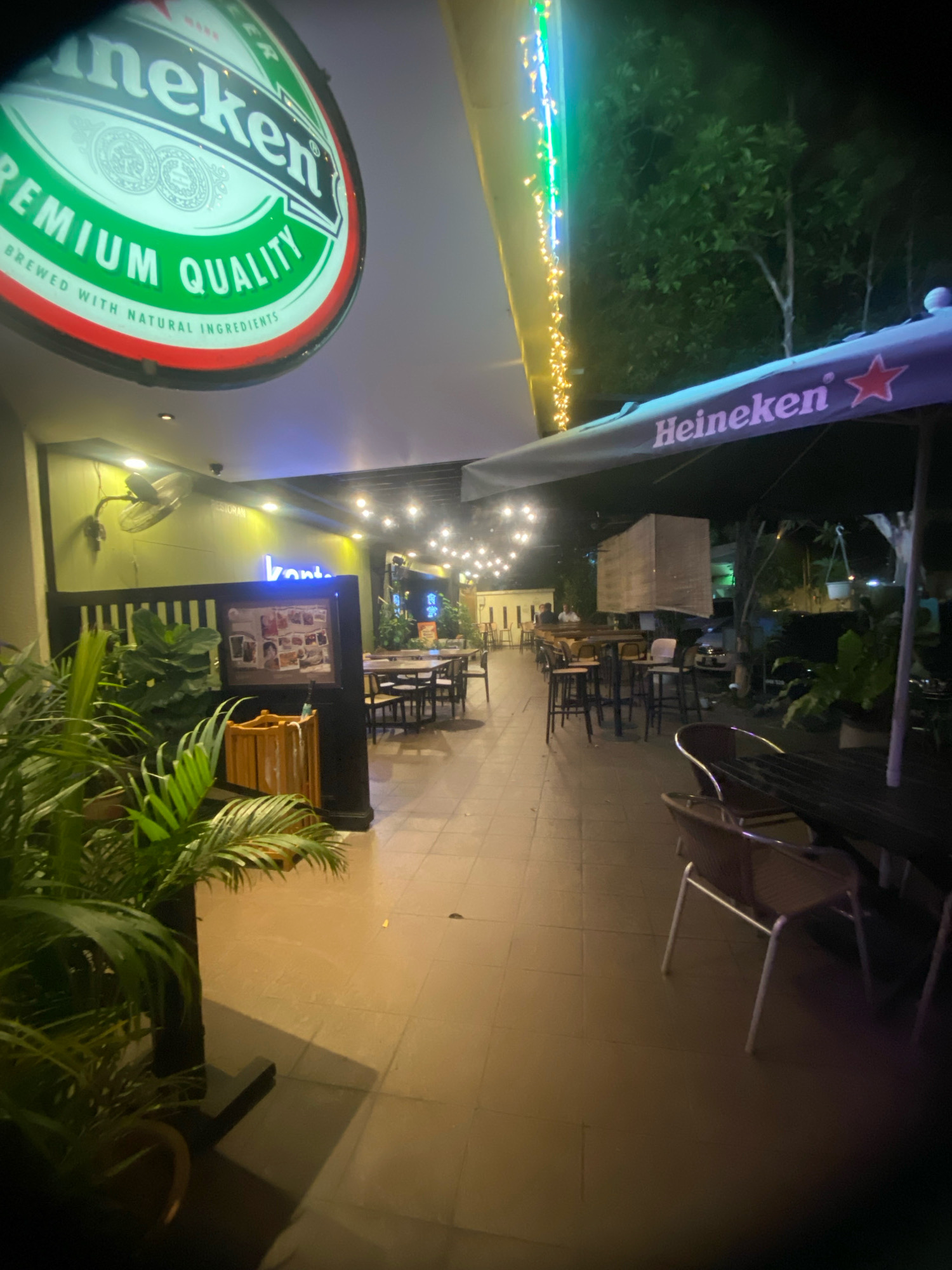 The image depicts an outdoor seating area of a restaurant or bar, likely in Kuala Lumpur, Malaysia, given the presence of Heineken branding, a popular brand there. The scene is illuminated by string lights and neon signs.
In the foreground, to the left, a large potted plant with lush green leaves is visible, partially obscuring the view of a wooden trash bin. Behind the plant, a dark partition with vertical slats separates this area from the rest of the establishment. A framed collage of photographs is mounted on this partition.
Further into the scene, an outdoor seating arrangement is laid out with numerous tables and chairs. Some tables are round with metal bases, while others appear to be rectangular. The chairs are a mix of styles, including some with metal frames and woven seats and backs, and others that are fully upholstered. In the background, there are more seating areas and the facade of a building with windows.
Overhead, a large Heineken umbrella provides shade, and a neon sign spelling "banten" is illuminated against a green wall. Further back, a string of fairy lights is draped across the area, casting a warm glow. The ground is paved with square tiles. In the distance, several trees are visible against a dark sky, suggesting it is nighttime. The overall impression is a relaxed, ambient evening setting for patrons.