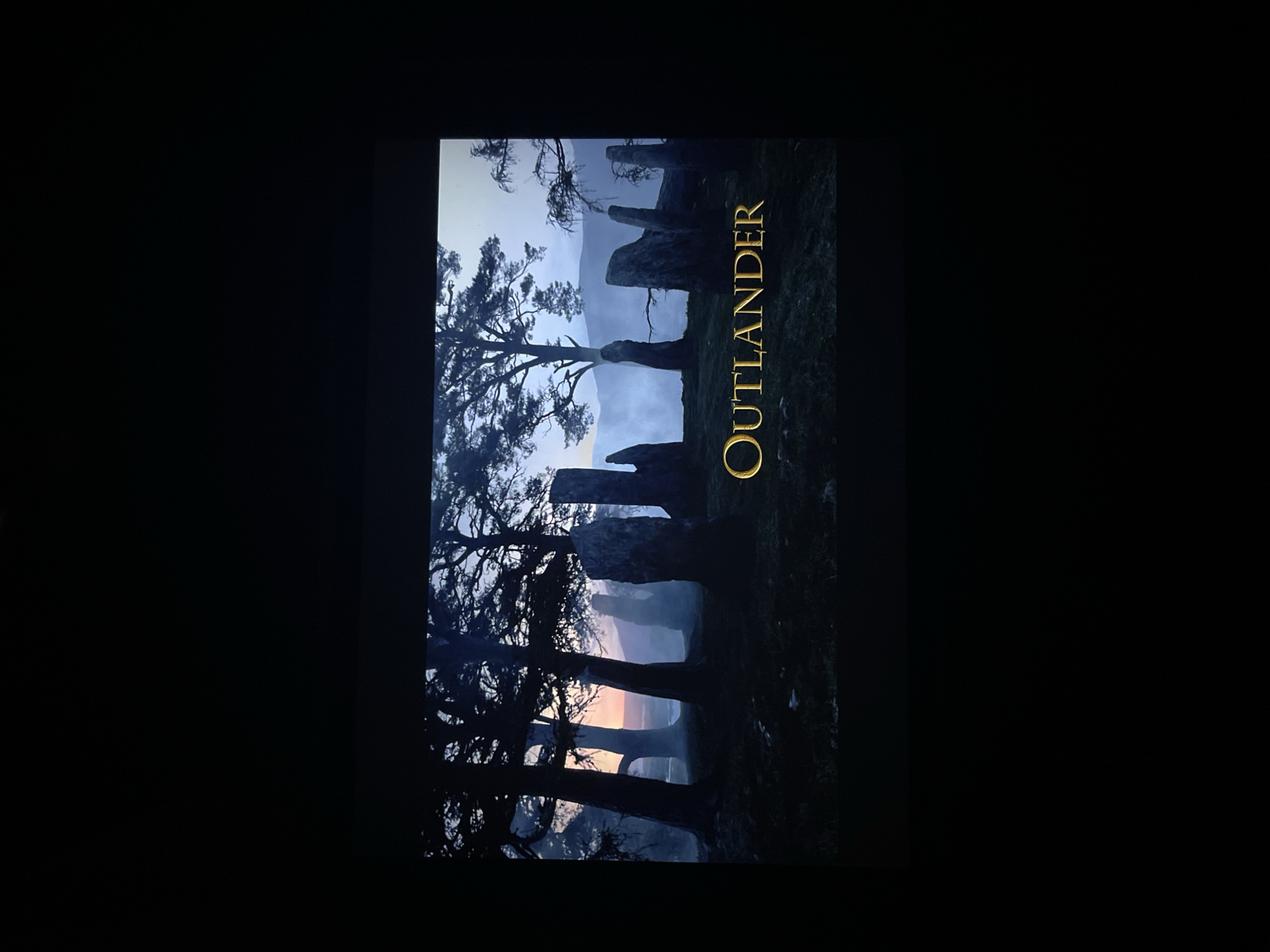 The image is a vertical movie poster for "Outlander." The poster features a landscape of standing stones and silhouetted trees against a backdrop of a hazy, twilight sky. The text "OUTLANDER" is written vertically in golden lettering on the right side of the image. The scene appears to be set in a rural or forested area, likely a historical setting. There is no indication that this image is in Varna, Bulgaria.