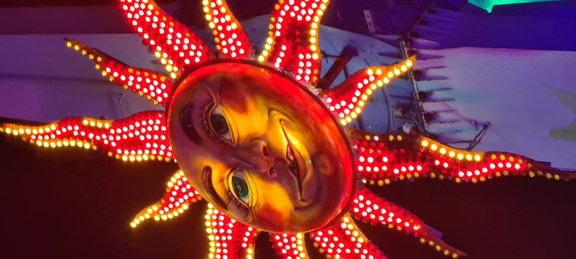 The image depicts a decorative sun-shaped fixture. The face is painted with realistic human features: eyes, nose, and a mouth. The sun's rays extend outward from the face and are illuminated with rows of yellow and red lights. The overall impression is that of a carnival or amusement park decoration. No people are visible. The specific location is unknown without further information. The environment is obscured and dimly lit, with some background elements suggesting a structure, possibly part of a carnival ride.