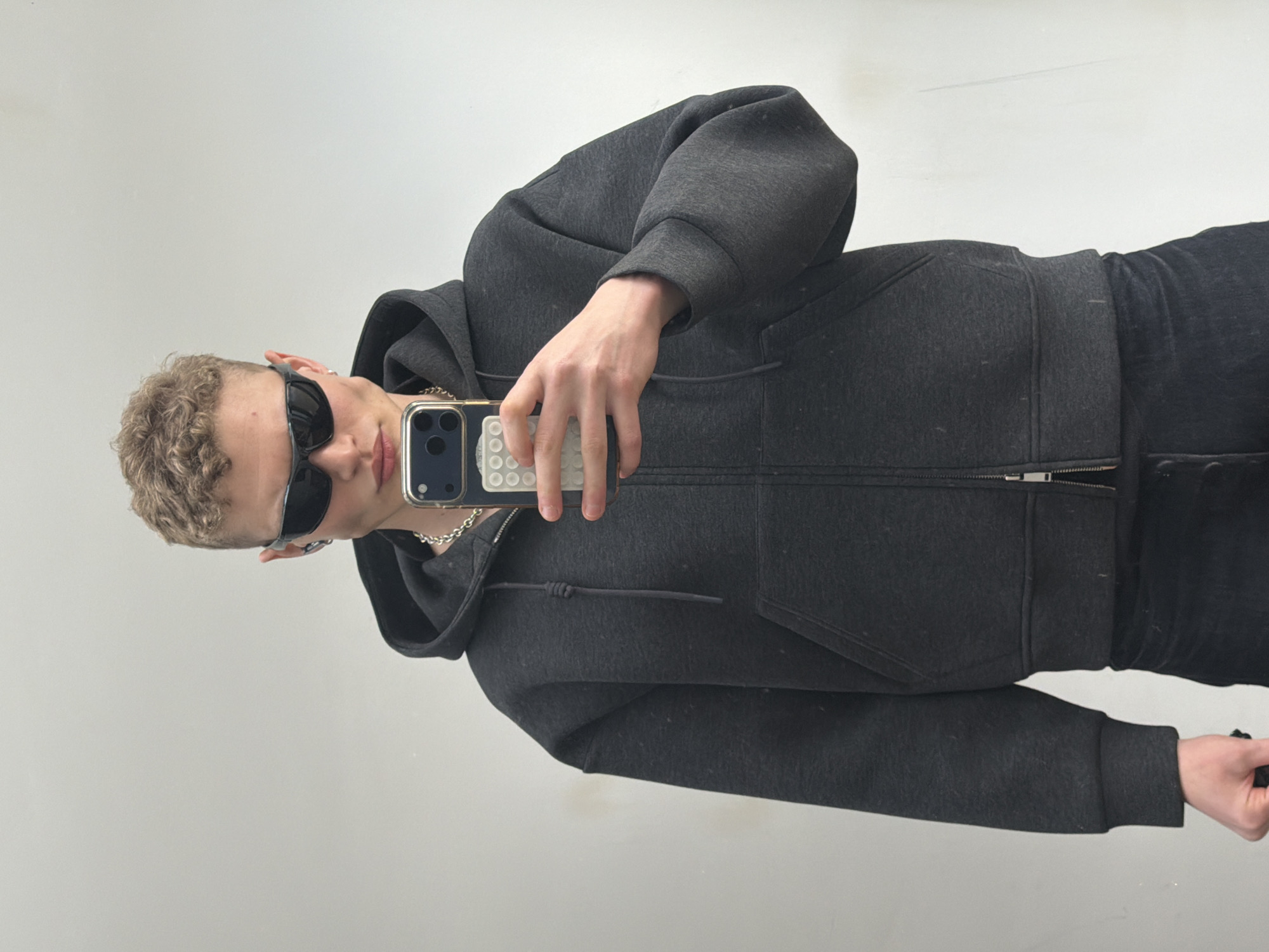 A person with short, curly blonde hair, wearing sunglasses and a dark grey hoodie, is holding a phone up to take a selfie in front of a plain, light-colored wall. The person is also wearing a silver chain around their neck and dark-colored pants. The phone has a white, textured case. The scene appears to be indoors, potentially a studio or a minimalist space. There is no indication of the location being London, United Kingdom.