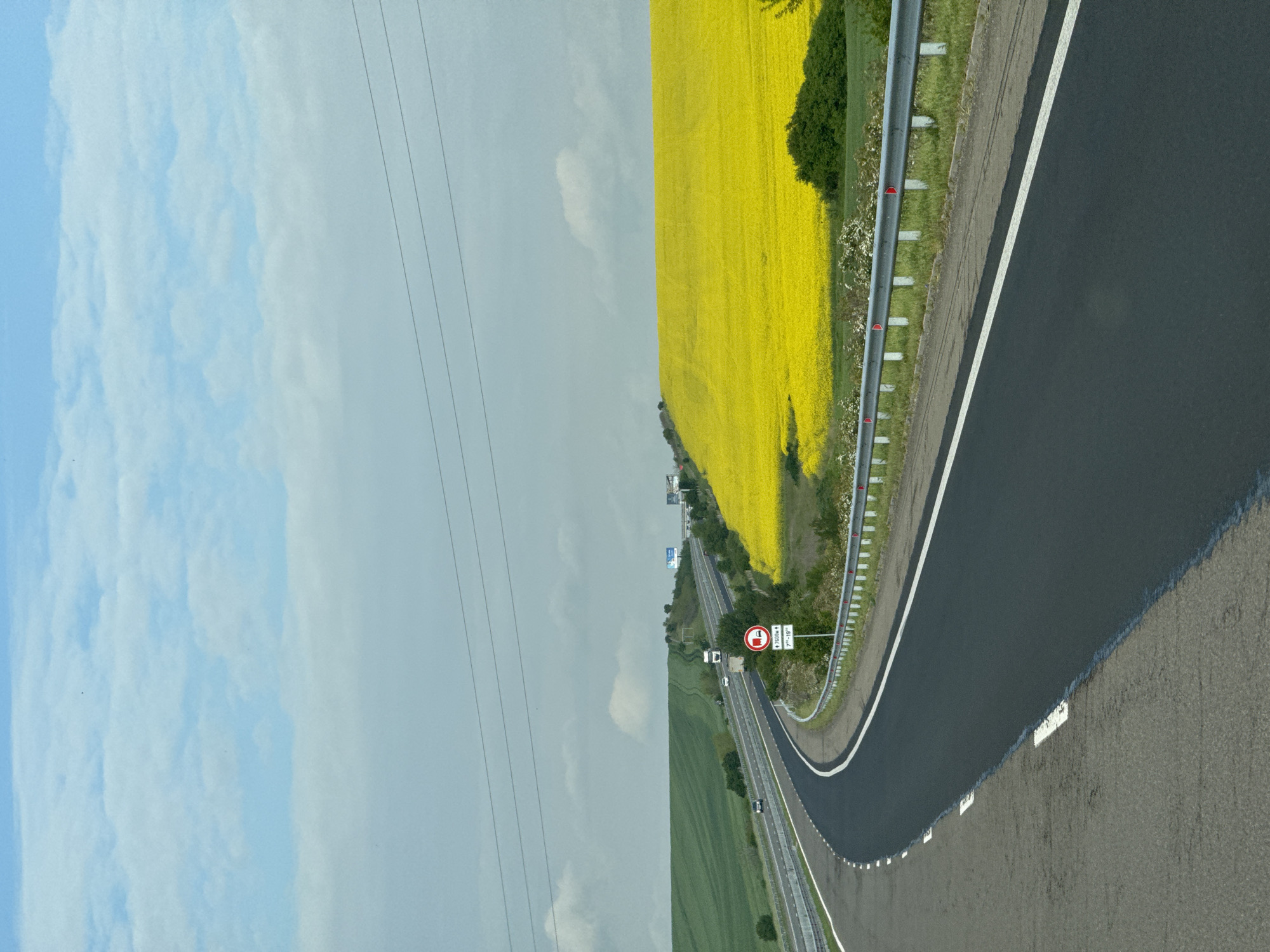 The image depicts a road scene. On the right, a field of yellow flowers borders the road. To the left, the sky transitions from blue with clouds to overcast. In the foreground, a highway curves to the right, marked with a white line and a guardrail. A road sign is visible adjacent to the road, featuring a red and white symbol and numbers, indicating a height or weight restriction. The road surface shows a contrast between a freshly paved dark section and an older, lighter gray section. Power lines stretch across the sky in the center. The scene suggests a rural setting with highway infrastructure. Based on the metadata and information available, this image is captured from or represents a location in the city of Troyanovo, Bulgaria.