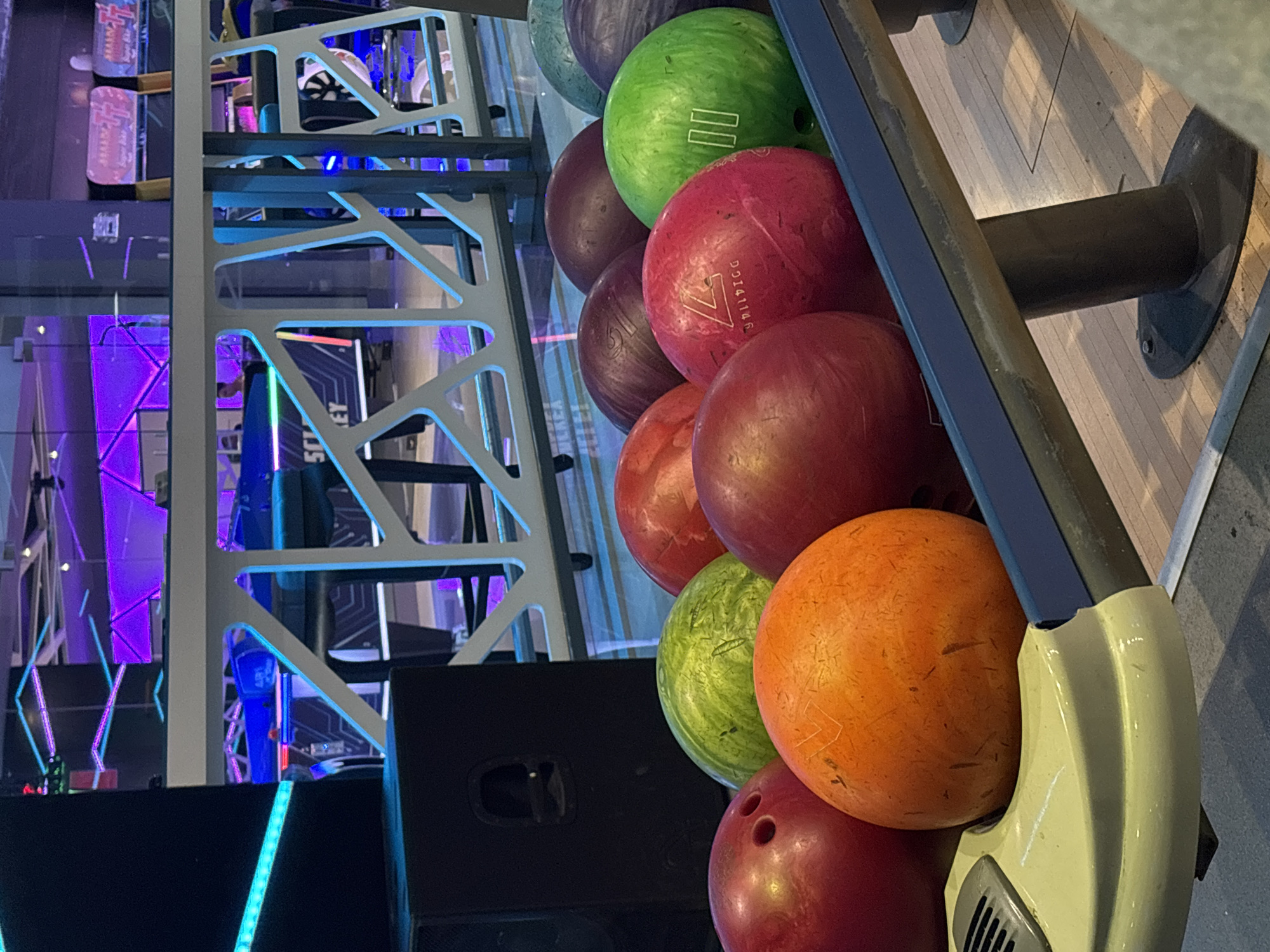The image depicts a selection of bowling balls in a rack, likely at a bowling alley, possibly in Gabrovo, Bulgaria, but not confirmable from the image. The bowling balls are of various colors, including green, red, purple, and orange. The bowling ball rack is gray and blue, and is mounted on a wooden platform. In the background, there are neon lights and structural elements, which may be a part of the entertainment venue. There are no people visible in the image.