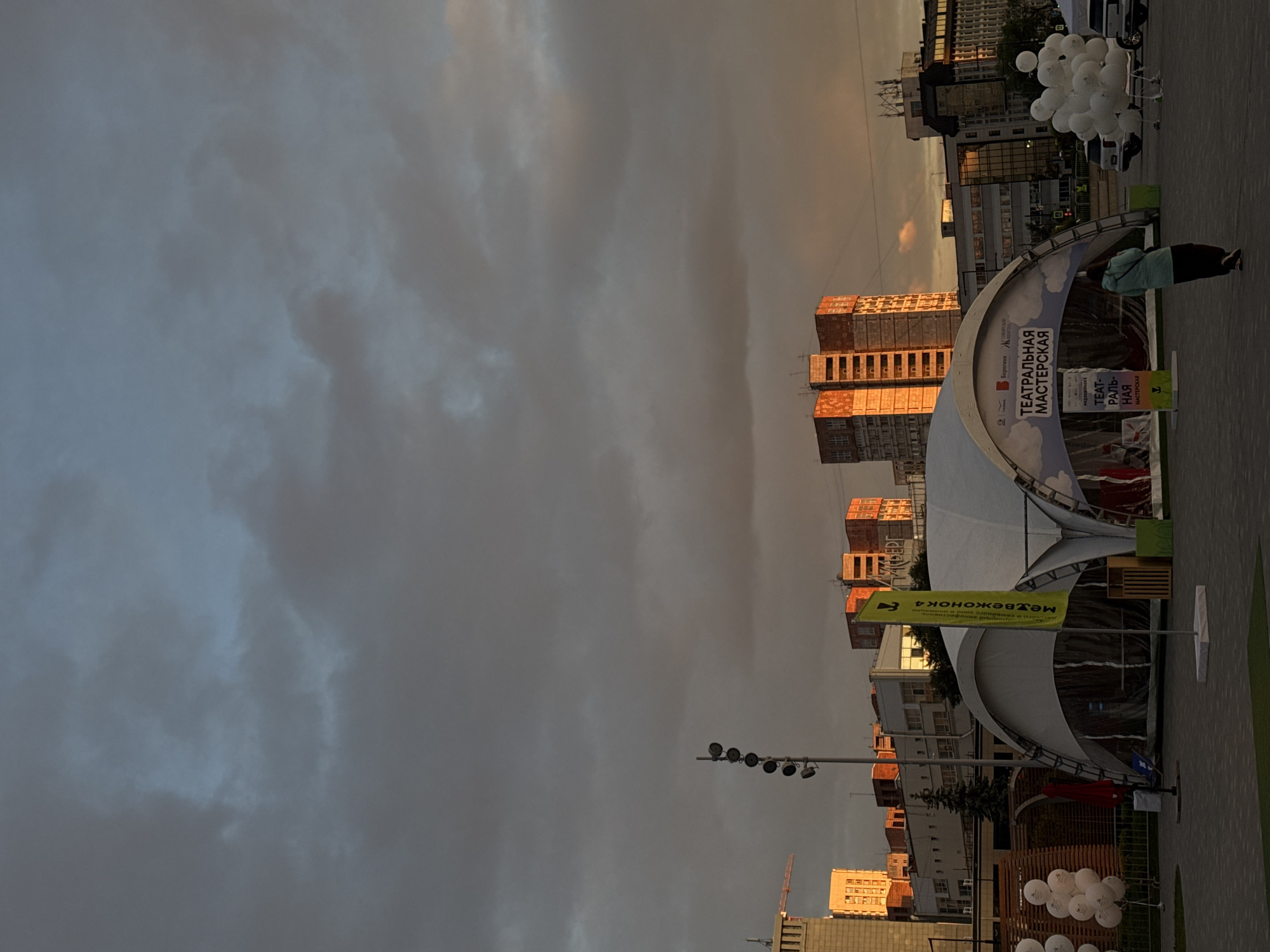 The image shows a cityscape in Perm, Russia, with a dramatic sky of gray and golden clouds. The scene includes buildings, particularly tall structures that catch the setting sun, creating a warm glow. A prominent feature is an outdoor event or festival, indicated by a large white tent structure. Text that can be read includes "TEATPAЛЬНАЯ МАСТЕРСКАЯ", which translates to "Theatrical Workshop", suggesting the event is related to performing arts. There are also balloons present. One person is visible near the tent. Street lights and other urban features are also visible.