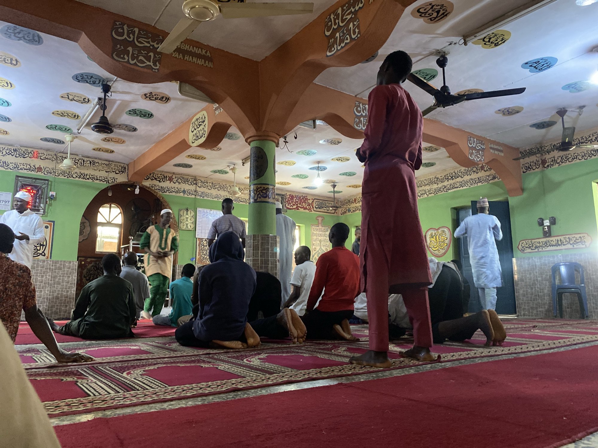 In an interior, potentially a mosque, a group of individuals are engaged in prayer. The scene is illuminated by overhead lighting and ceiling fans. Religious inscriptions adorn the ceiling and walls. A man in red clothing stands observing the praying individuals. The floor is covered with a red carpet. It's indicated by text in the image that this location is in Lagos, Nigeria.