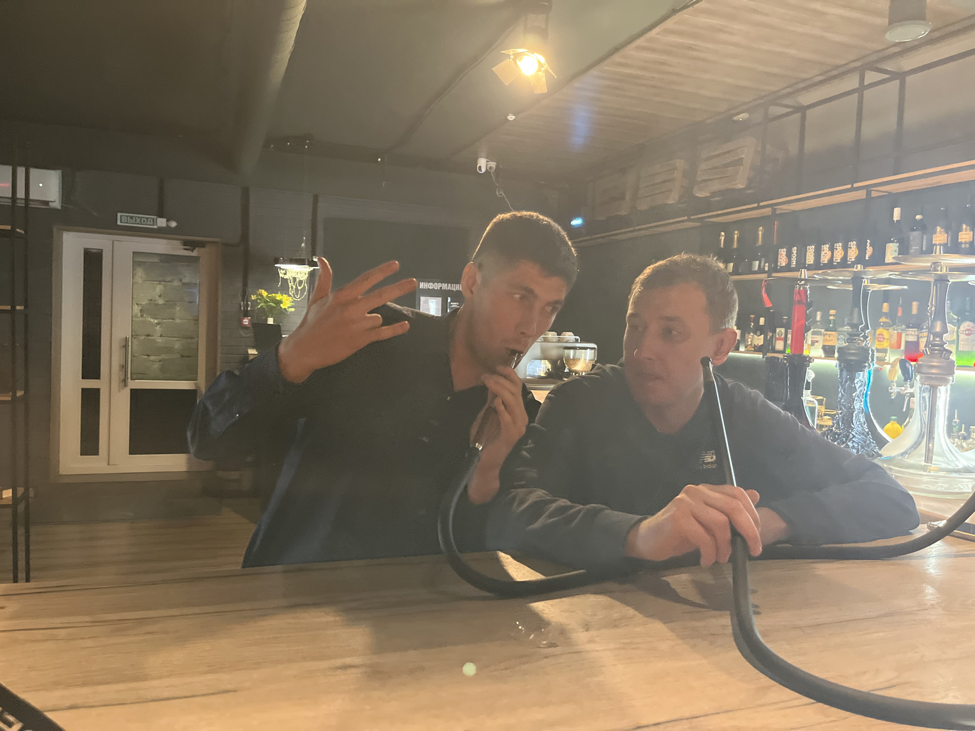Two adult males are seated at a light-colored wooden bar or high table within a dimly lit commercial establishment, likely a lounge or hookah bar, located in Chaikovsky, Russia. The male on the left, wearing a dark long-sleeved shirt, is actively engaged in conversation, gesturing with his raised left hand, exhibiting an extended thumb, index, and pinky finger while his middle and ring fingers are bent. His right hand holds a black hookah hose near his mouth. The male on the right, also in a dark long-sleeved shirt with a white "New Balance" logo visible on his left sleeve, is attentively listening, with both hands resting on the table, holding another section of a black hookah hose. A multi-hose hookah apparatus is prominently displayed on the bar surface to the right of the two individuals. The background features industrial-style ceilings with exposed elements and several spotlights providing illumination. Behind the individuals, liquor bottles are arranged on dark-framed shelves, typical of a bar setting. To the far left of the frame, a white door is visible, above which a green illuminated sign displays the Cyrillic word "ВЫХОД" (EXIT). Adjacent to the exit sign, a small security camera is mounted. Above the left male's head, another sign reads "ИНФОРМАЦИЯ" (INFORMATION).