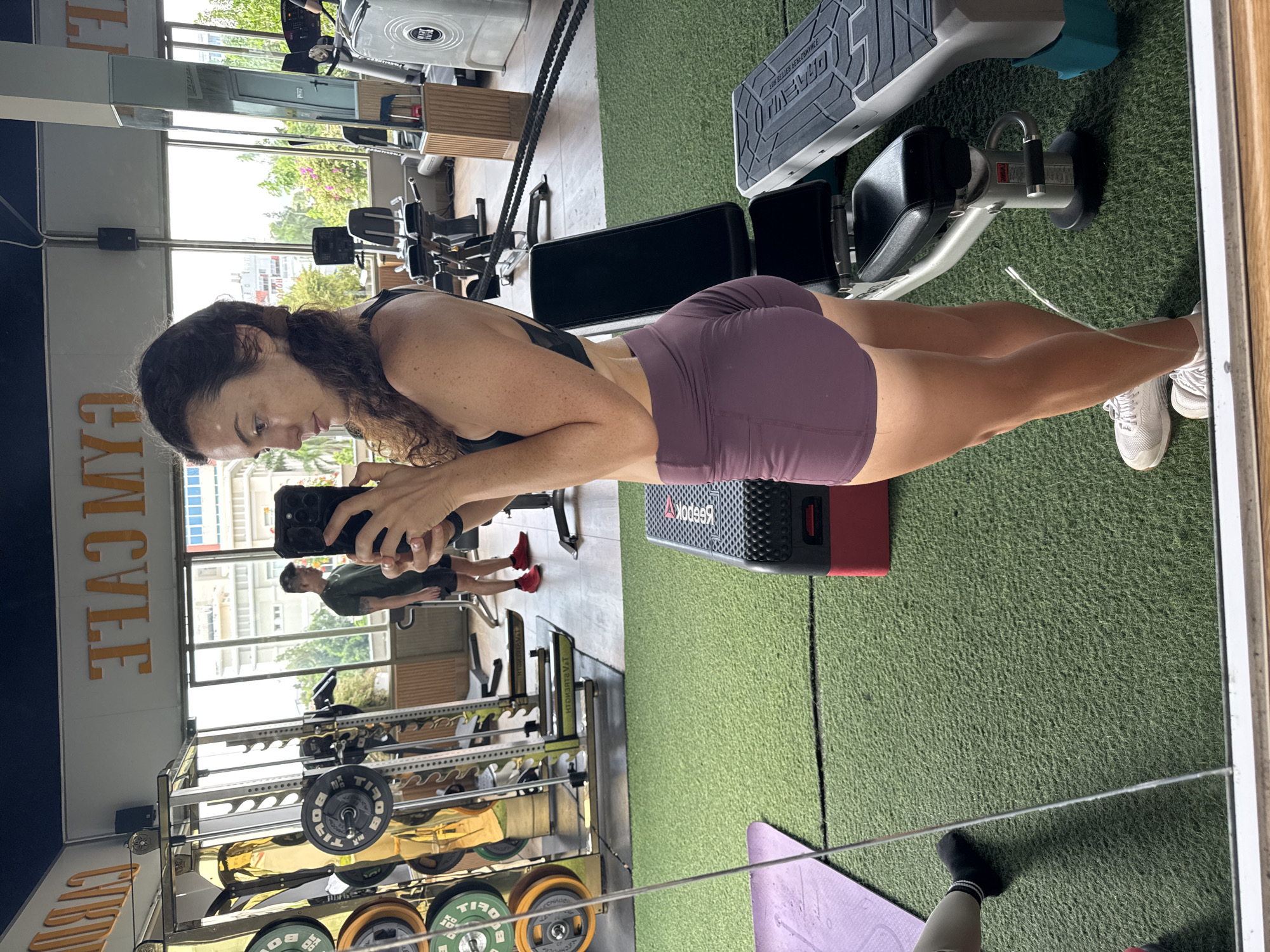 In a gym in Phan Thiết, Vietnam, a woman in a black sports bra and purple shorts takes a mirror selfie. She is holding a black smartphone. In the background, a man in a green t-shirt and black shorts stands near weightlifting equipment. The gym has artificial turf flooring, treadmills, weight racks, and benches. The gym features large windows offering a view of the city and greenery. The wall displays the word "GYM" in large orange lettering.