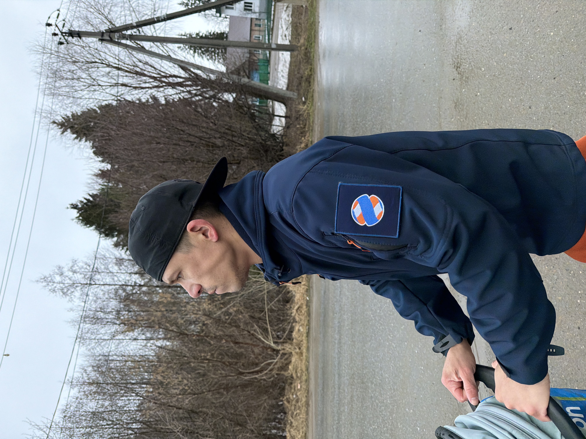 A man wearing a black baseball cap and a blue jacket with an insignia on the left sleeve is standing on a paved road in the city of Chaikovsky, Russia. He is bent over and appears to be holding the handle of a stroller. Behind him, there is a landscape with trees, some bare, and a house in the distance. Power lines are visible above the trees. The weather appears overcast, and the road looks wet.