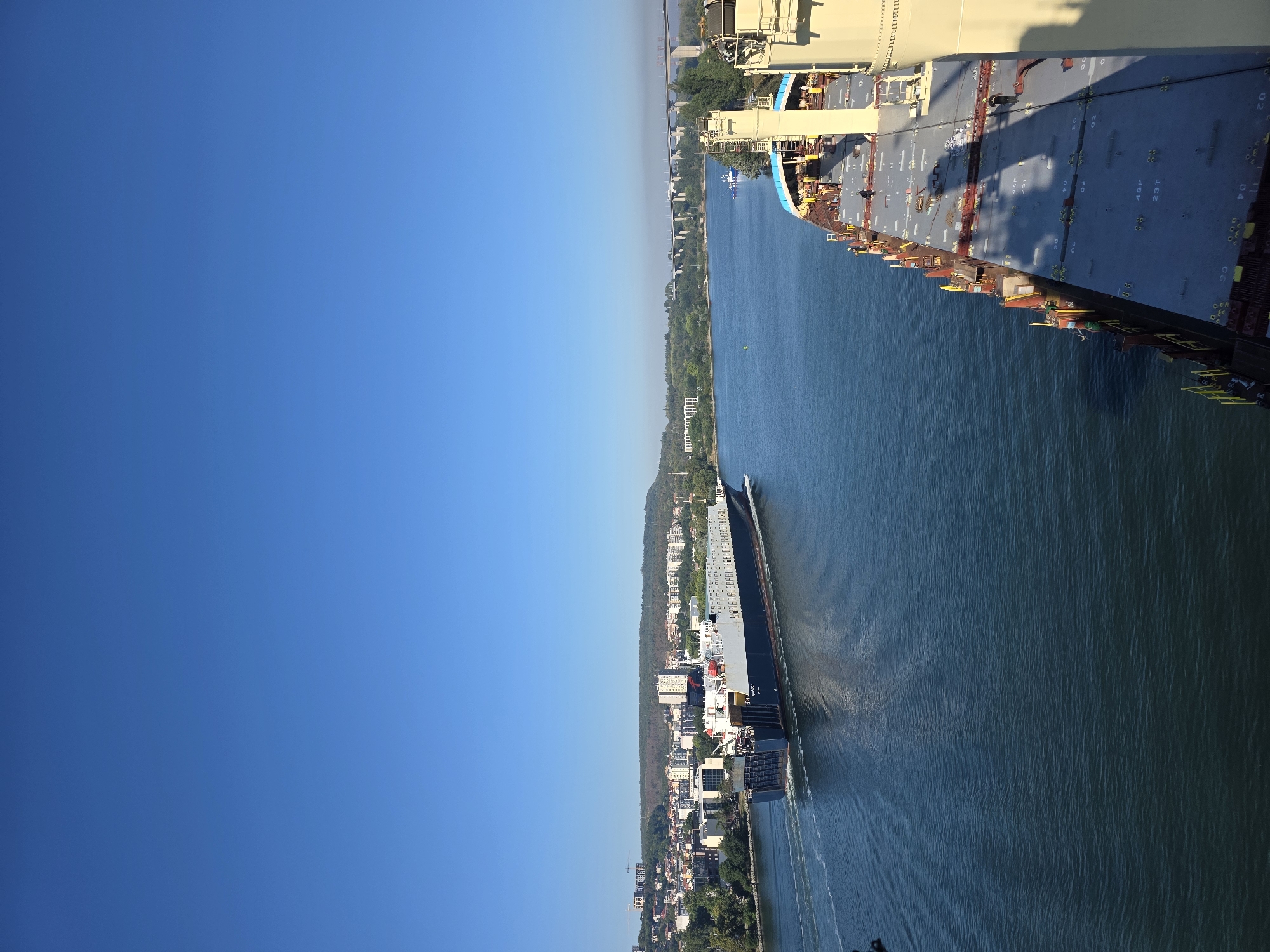 The image depicts a wide, clear blue sky over a bright, sunny day. Below the sky, a body of water, likely a canal or bay, is visible. On the left side of the water, a large cargo ship is traveling away from the viewer, leaving a wake behind it. The ship is dark blue on its lower hull with a white superstructure. The ship's name, "NAPOLI", is visible on its side. Along the far shore, a cityscape with buildings of various sizes and architectural styles is situated, backed by a tree-covered hill.
In the foreground, on the right side of the image, the deck of another large ship is visible. This ship has a predominantly gray deck with yellow and red accents on its railings and structures. Several figures, appearing to be crew members, are visible on the deck of this ship. The deck is covered in cargo hatches and other equipment. In the distance, across the water, a long, multi-span bridge can be seen, connecting two parts of the landscape. The scene suggests a bustling port or waterway in the city of Varna, Bulgaria.