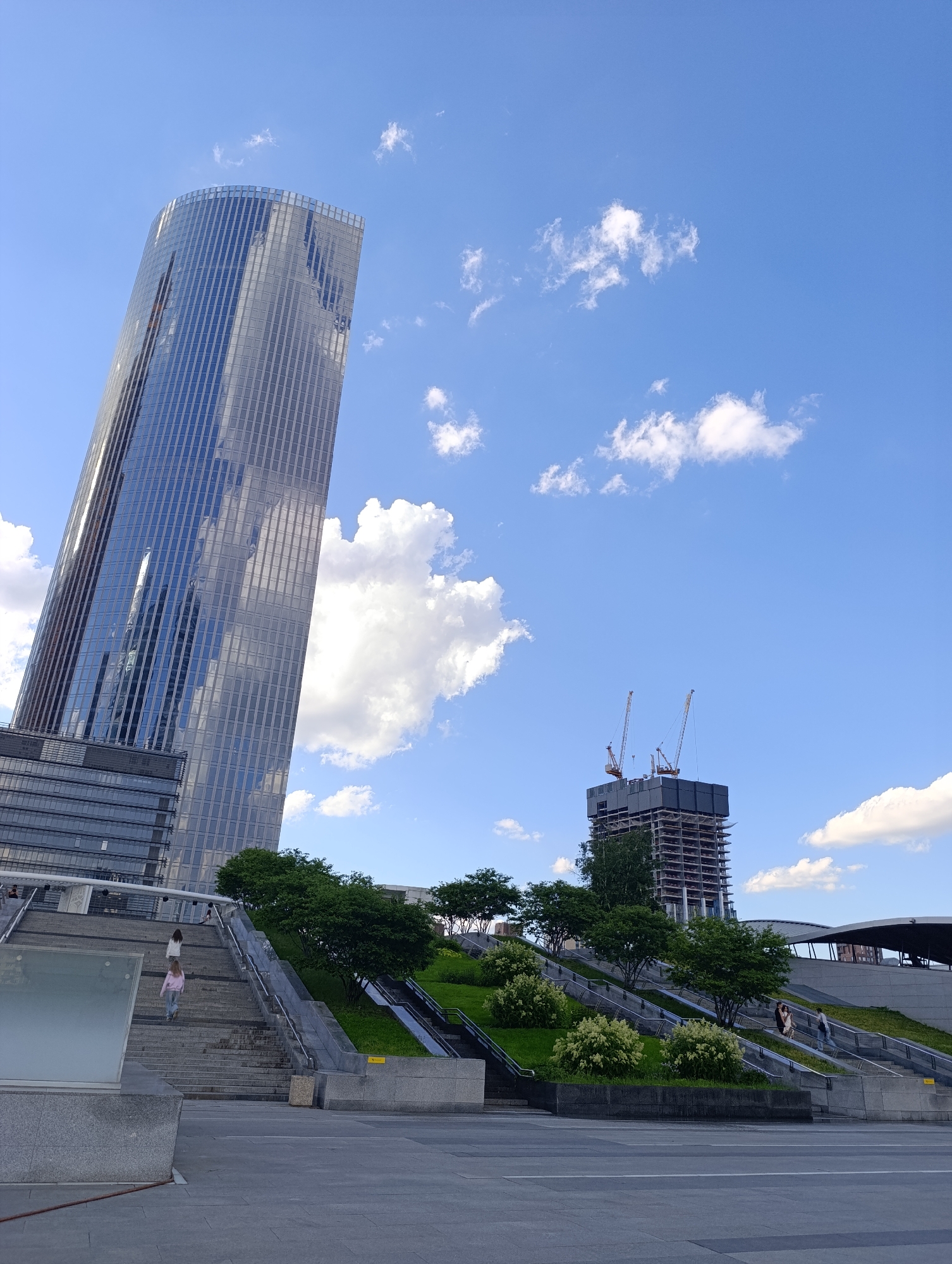 This vertical shot captures a modern urban landscape in Moscow, Russia, under a bright blue sky with scattered white clouds. The scene is dominated by a tall, slender, glass-clad skyscraper on the left, its curved facade reflecting the sky and clouds. Its sleek design features numerous vertical lines from its glass panels.
To the right, in the mid-ground, a building under construction stands, showing its reinforced concrete structure and topped with two yellow construction cranes. Below and in front of these buildings, a multi-tiered public space unfolds, featuring wide concrete staircases ascending a gentle slope. These stair sections are interspersed with grassy embankments planted with small trees and white flowering bushes, providing a touch of green to the urban environment. Escalators are also visible alongside some of the stairways.
Several people are present, indicating routine pedestrian activity. On the left-hand staircase, two women, one in a light pink top and jeans, and another in a white top, are walking upwards. Further to the right and higher up, a small group of people can be seen ascending another section of stairs. The bright daylight and clear shadows suggest it is daytime, likely a sunny morning or afternoon. The overall impression is one of a bustling, modern city with ongoing development and well-maintained public spaces. No discernible text is visible.