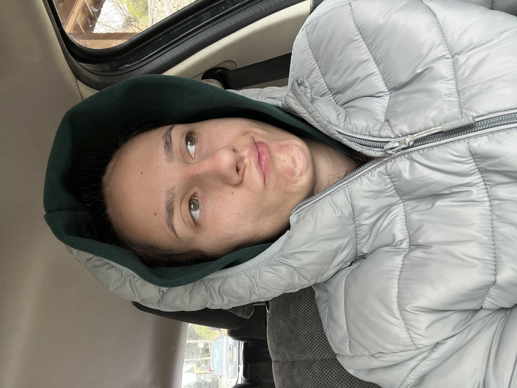 A light-skinned woman with brown hair is lying in the backseat of a car, wearing a light gray puffer jacket with a dark green hood. The label "PULL&BEAR" is visible on the jacket. The car's interior is visible, including the ceiling, a safety handle, and the car seat. Through the car window, blurry objects suggest a natural environment outside with what appears to be a wooden structure with a window. The scene appears to be Hrishteni, Bulgaria, as per the prompt.