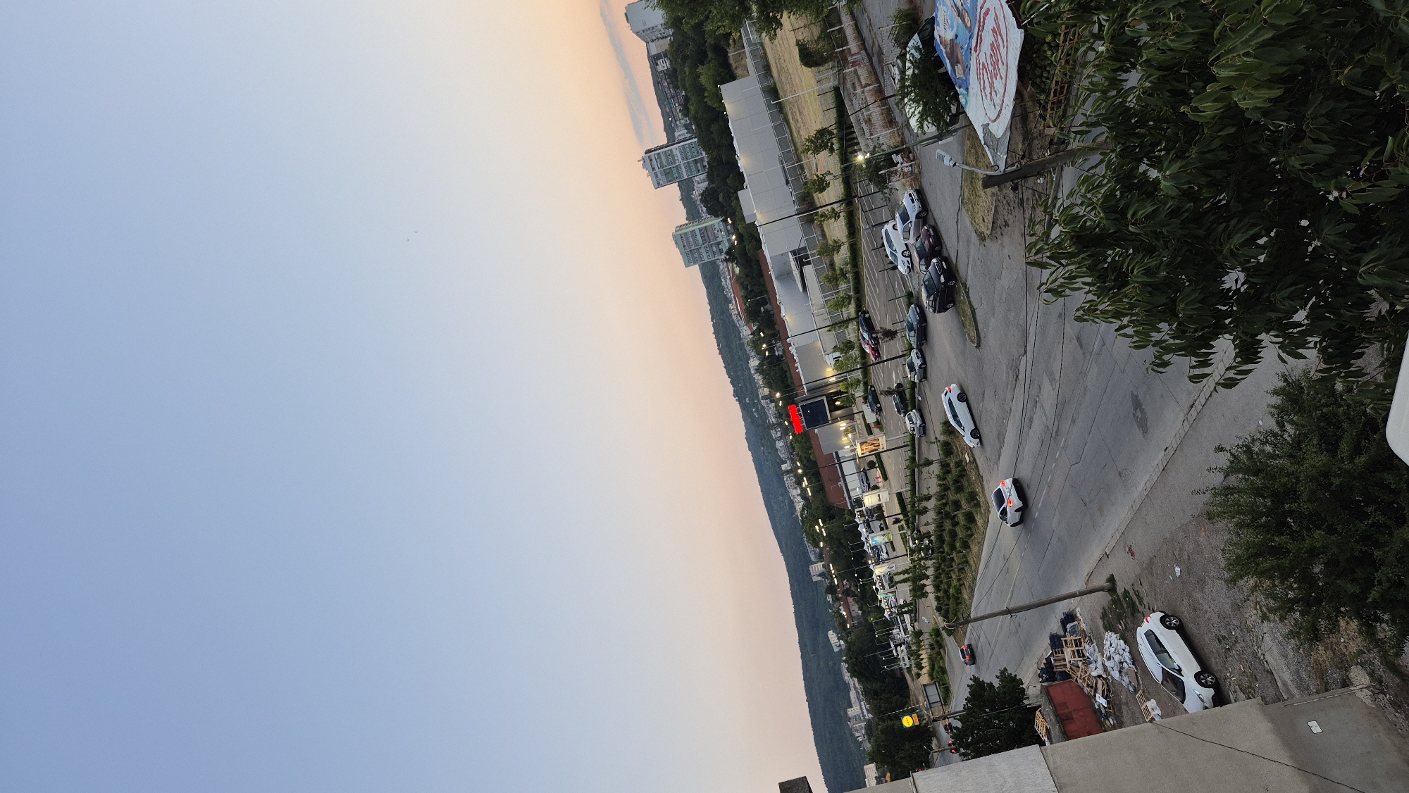 An elevated, wide-angle view captures a cityscape at dusk. The sky transitions from a clear blue at the top to a soft orange and pink hue near the horizon. A gently rolling, tree-covered hill forms the backdrop of the scene. In the middle ground, a commercial area features a large building with a red sign, likely a supermarket or retail store, with a parking lot filled with various cars. Several other buildings, including multi-story apartment complexes, are scattered throughout the city. In the foreground, a two-lane road curves through the scene, with several cars driving on it. A white car is seen from behind in the foreground, with another white car parked on the left side of the road. Trees and bushes are present throughout the urban landscape, with lush green foliage visible in the lower right and left portions of the image.