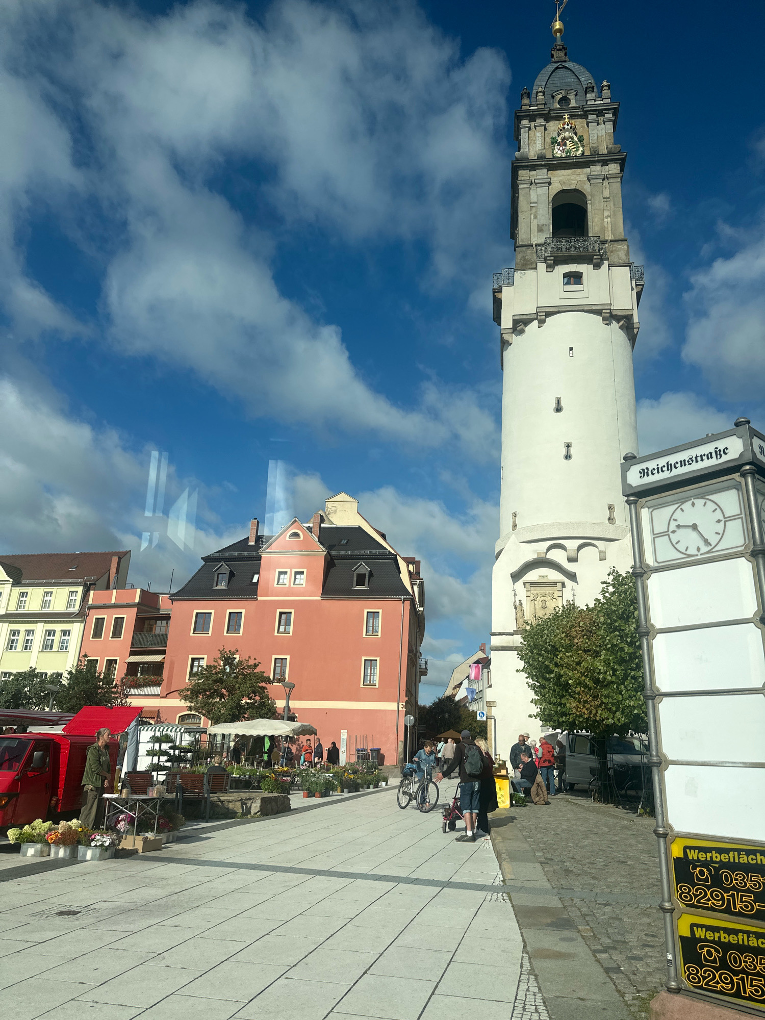 The image shows a street scene in Bautzen, Germany, focusing on the Reichenturm (Reichenbach Tower). A tall, white tower with a clock face and a dark spire dominates the right side of the frame. In front of the tower, a street clock with the inscription "Reichenstraße" is visible. To the left of the tower, several colorful buildings are visible, including a prominent salmon-pink building. A market is set up in the foreground with stalls selling flowers and plants. People are seen browsing the market, walking, and cycling. A red van is parked near one of the stalls. The sky is blue with scattered white clouds. The ground is paved with light-colored tiles. The overall impression is of a bustling town square on a sunny day.