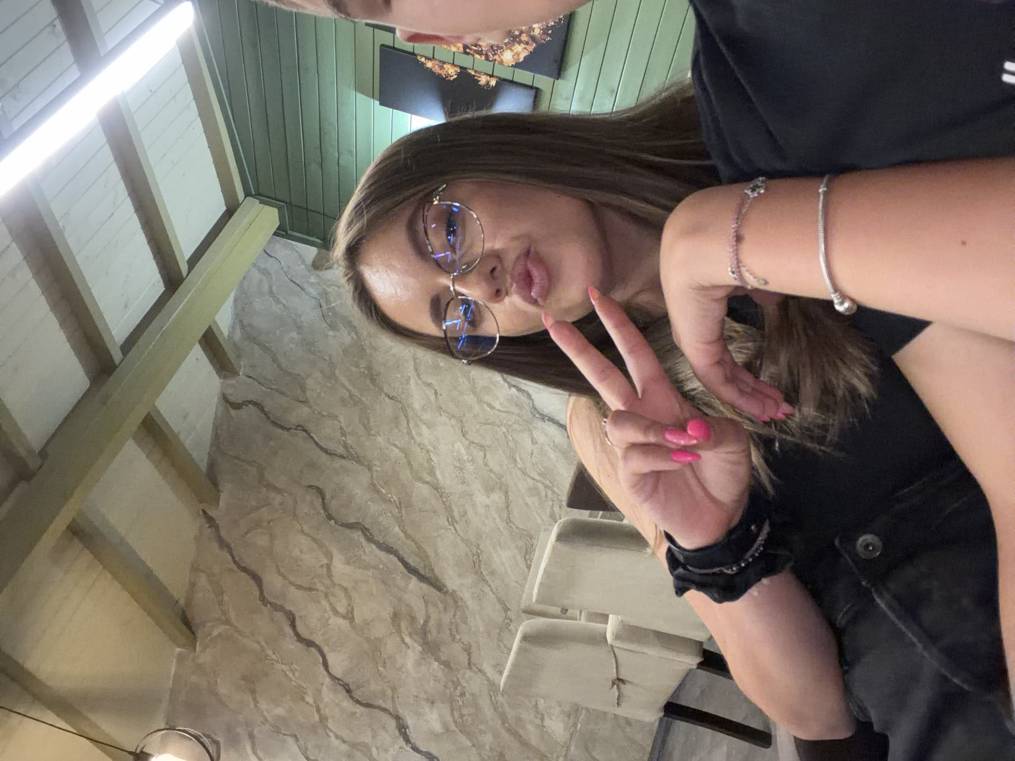 A woman, wearing glasses, a black top, and denim jacket, poses for a selfie, puckering her lips and making a peace sign with her fingers. She has bright pink fingernails and wears two bracelets on her left arm. Behind her, two chairs are visible, and the wall features a textured, marbled pattern. The ceiling is made of wooden beams and white panels. The scene appears to be indoors, possibly in a restaurant or cafe. There is no visual information in the image that links it to the city of Panagyurishte, Bulgaria.