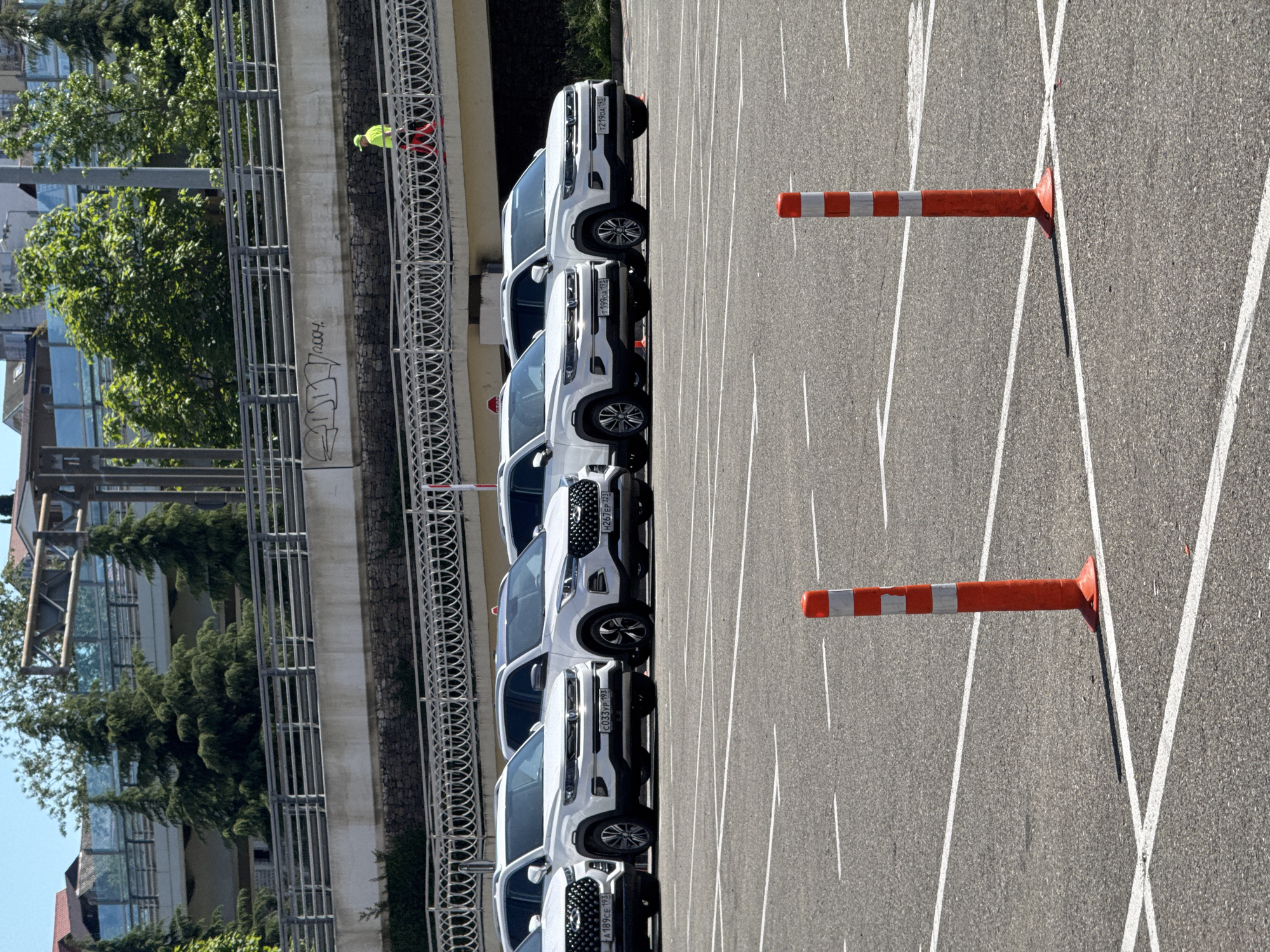 The image captures an outdoor scene, likely a parking lot or driving course, under bright daylight conditions. A line of at least six identical white SUVs, possibly Chery Tiggo 7 Pro models, are parked facing forward on asphalt pavement marked with white lines.
On the far left, a tall concrete barrier or wall runs parallel to the parked cars, topped with a metal fence. Behind this barrier, lush green trees and glimpses of buildings are visible. One person is present on the concrete structure, wearing a bright yellow high-visibility jacket and red clothing, appearing to observe the area.
Two orange and white striped traffic cones or barriers lie horizontally on the asphalt in the foreground and mid-ground, suggesting an organized setup such as a driving school, vehicle testing area, or temporary parking zone.
Notable details include the sunny weather, indicated by strong shadows. Visible text includes several Russian license plates on the parked cars: "T219OA 193", "T199OA 193", "H261EP 123", "C033TP 193", and "A189CE 193". The "193" and "123" region codes correspond to Krasnodar Krai, where Sochi is located. Additionally, faint graffiti is visible on the concrete wall, including what appears to be "2007" or "2004" and some indistinct letters.