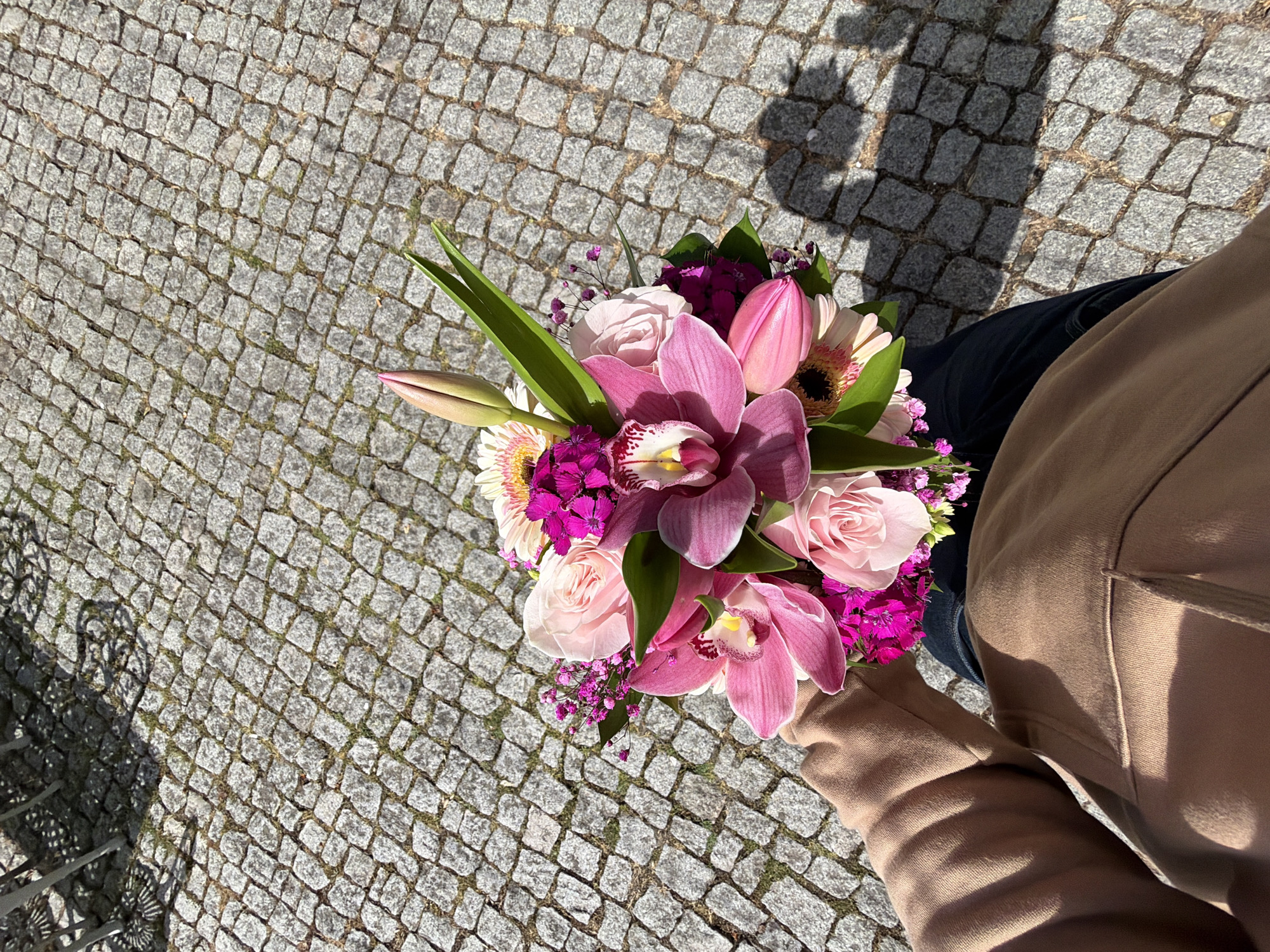 A person holds a bouquet of flowers over a cobbled pavement. The bouquet features pink orchids, roses, tulips, gerberas, and small purple flowers. The person is wearing a tan-colored jacket and dark jeans. The scene is outdoors, with the cobbles suggesting a location in an urban area, possibly in Vila Real, Portugal, given the user's provided context. The shadow of the person holding the flowers is visible on the pavement.