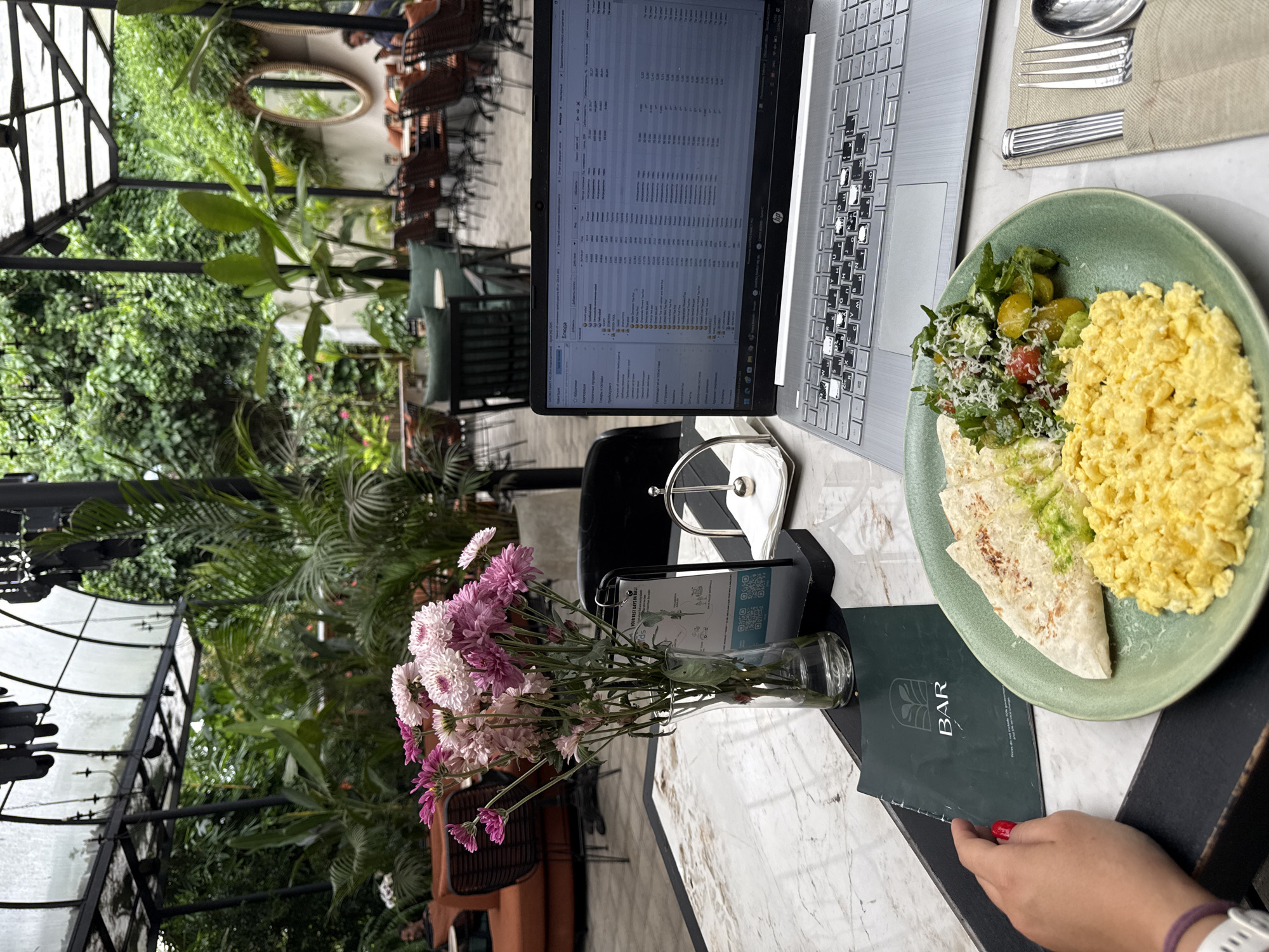 The image shows a laptop and a plate of food on a table, possibly in Ubud District, Indonesia. A laptop displays a spreadsheet or similar data-heavy application. To the right, a green plate contains scrambled eggs, a salad with tomatoes and avocado, and what appears to be a folded flatbread. Silver cutlery wrapped in cloth sits near the plate. To the left, a glass vase holds a bouquet of pink and white flowers. A menu card with the word "BAR" is visible below the plate. A person's hand with red nail polish is holding the edge of the menu card. The background reveals an outdoor or partially covered patio with lush greenery. The furniture indicates a relaxed, cafe-style setting.