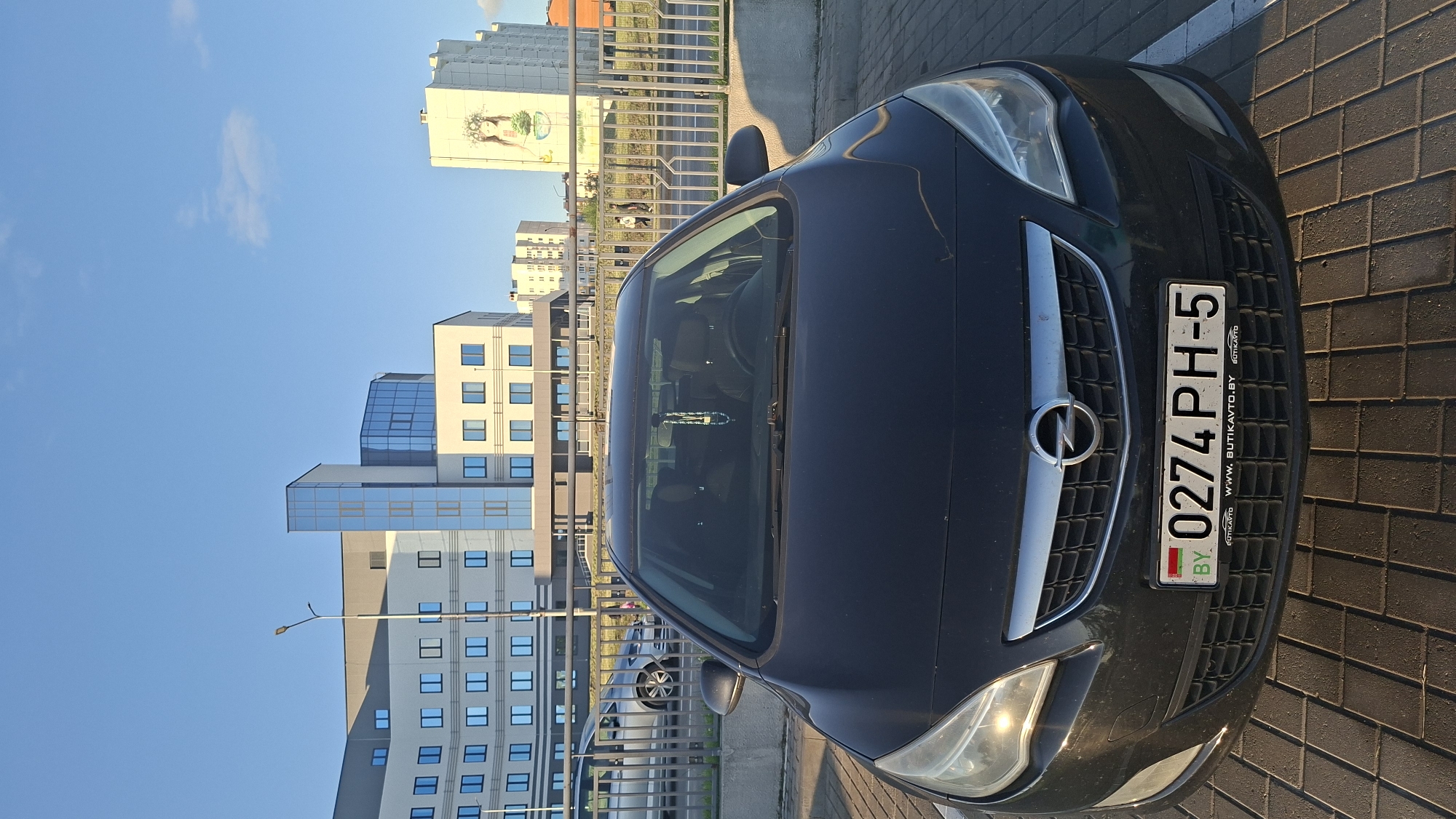 In Smaliavichy, Belarus, a black Opel Astra is parked on a paved lot. The car's license plate reads "0274 PH-5" and features the BY country code. In the background, a modern building with a glass facade stands against a clear blue sky. Further in the distance, another building displays a mural of a woman adorned with a floral wreath. A metal fence separates the parking area from the street.