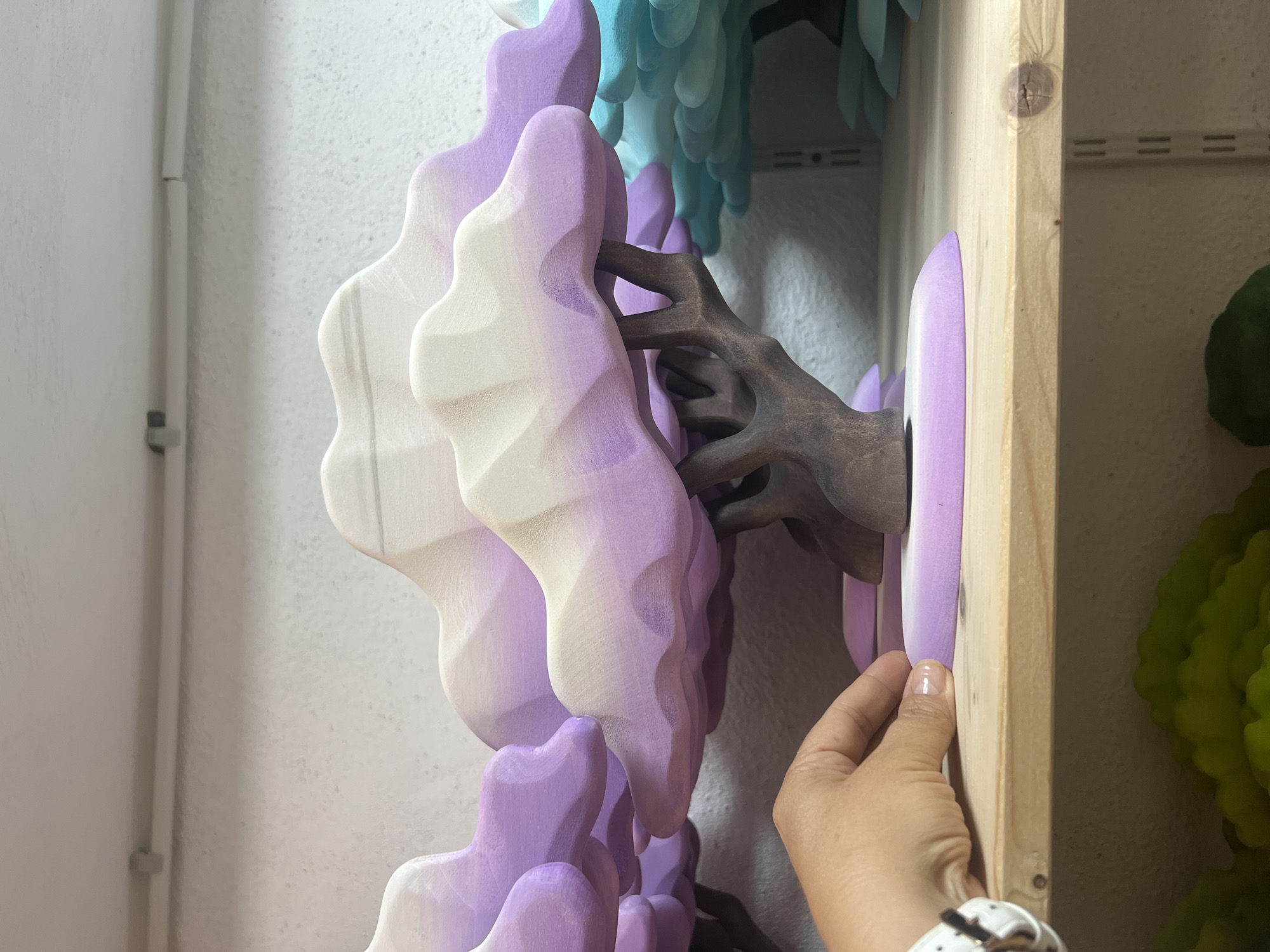 A hand with a watch on its wrist is shown in the foreground, touching the base of a wooden decorative tree on a shelf. The tree has a brown trunk and branches, and its canopy is made of layered, rounded shapes painted in a gradient of white to purple. The shelf is made of light-colored wood and is mounted on a wall with adjustable shelf brackets visible below. In the background, other similar decorative trees in shades of blue and green are partially visible. The overall setting appears to be an indoor display or storage area.