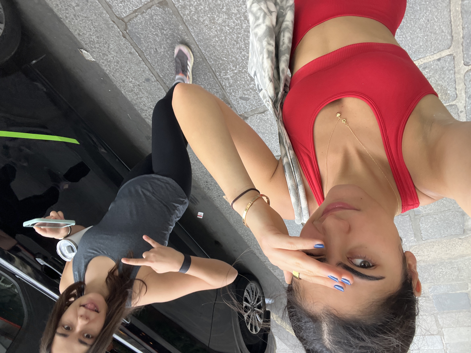 Two young women are visible in this outdoor, street-level image. The woman on the left is closer to the camera, taking a selfie. She has dark hair pulled back and is wearing a red sports bra and red shorts. She has blue nail polish and is touching her forehead with her right hand. She is also wearing a gold necklace and a gold ring. A grey and white patterned bag is slung over her left shoulder.
The woman on the right is further back and to the right. She has long, dark brown hair and is wearing a grey tank top and black leggings. She is making a "rock on" hand gesture with her left hand and sticking her tongue out. She is holding a white cylindrical object in her right hand and a light blue smartphone in her left.
They are standing on a grey cobblestone sidewalk next to a black vehicle. A portion of the vehicle's silver wheel is visible. The overall lighting suggests it is daytime, possibly overcast, as there are no strong shadows. The environment appears to be an urban setting. No specific landmarks are identifiable that would confirm the location as Paris, France, but the street and vehicle are consistent with a European city.