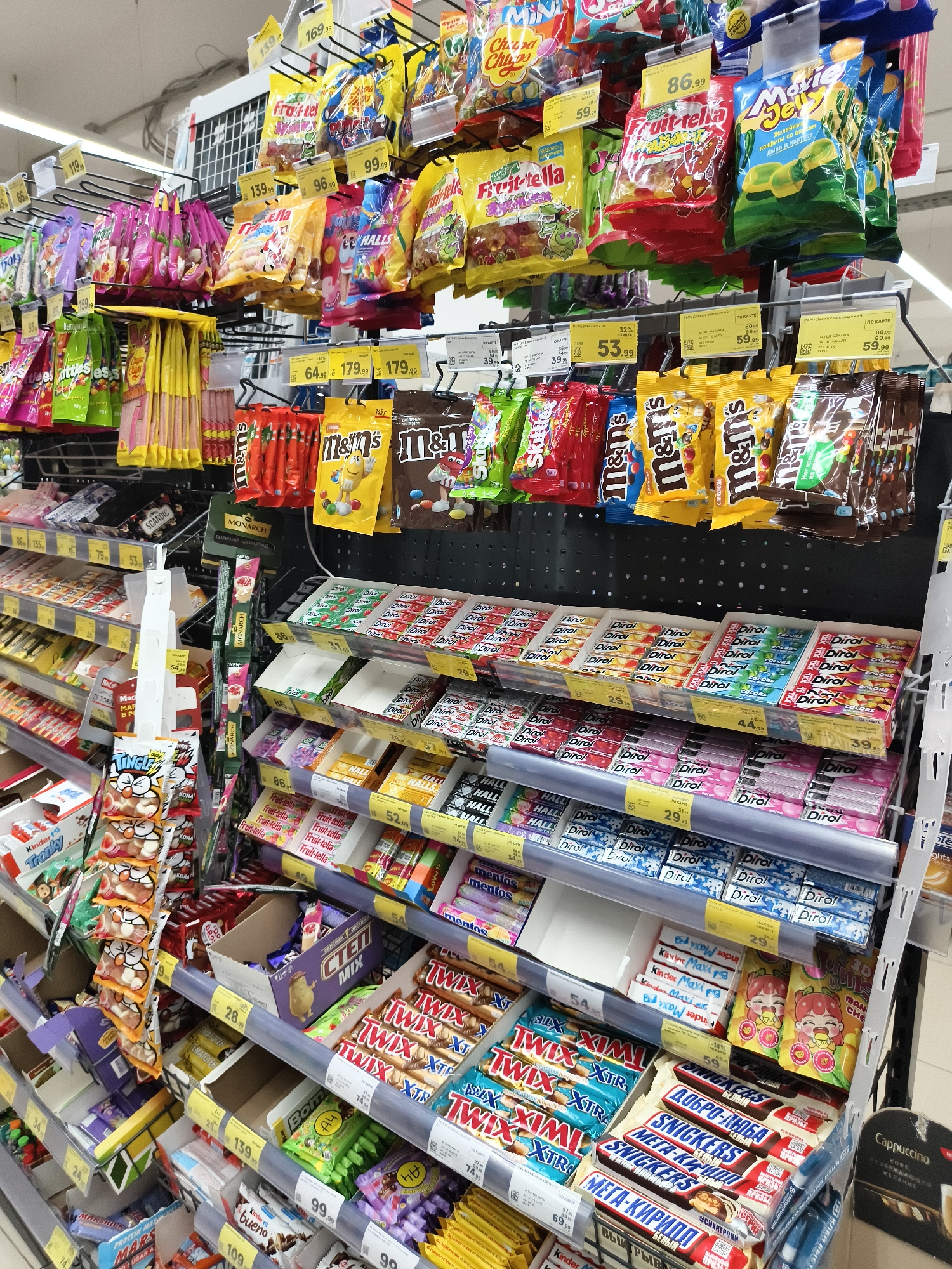 The image shows a brightly lit supermarket aisle with shelves stocked with various confectionery items, including candy, chocolate bars, and gum. The products are neatly arranged on metal and plastic shelving units. Prominent brands visible include M&M's, Twix, Snickers, Kinder, and Dirol. Many items have price tags attached. The environment appears to be a typical retail setting. The photograph was taken on August 15, 2023, in Chaikovsky, Russia. There are no people visible in the frame.