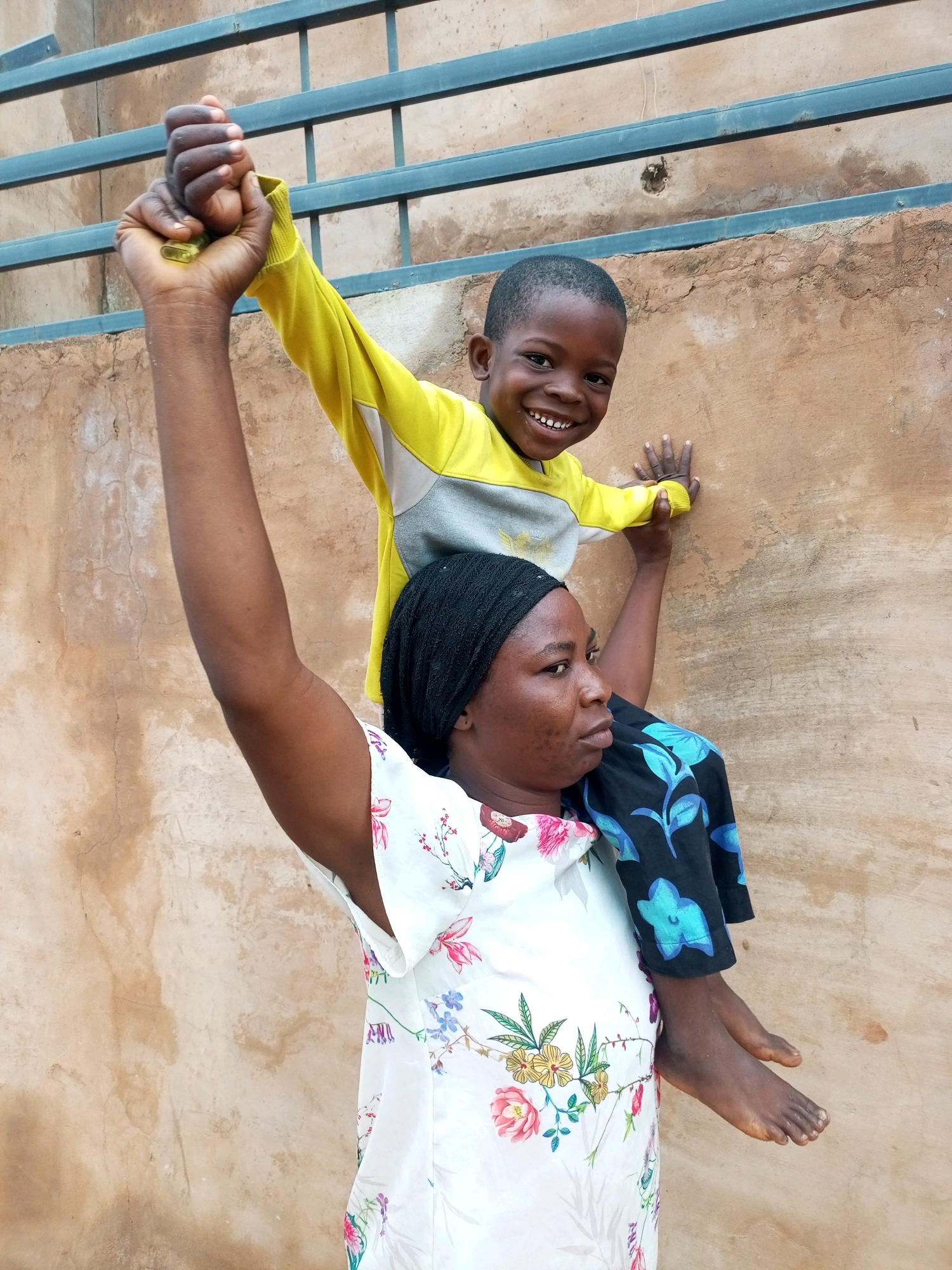 In the city of Tempure, Nigeria, a woman with dark skin and braided hair, wearing a floral-patterned dress and a black headscarf, carries a young boy on her shoulders. The boy, who appears to be of African descent, is wearing a yellow and grey long-sleeved shirt and black pants with blue floral patterns. He smiles at the camera, with his right arm raised and fist clenched, holding onto the woman's arm. The woman is looking slightly to her right, with a neutral expression. They are positioned in front of a rough, textured wall. Above them, a metal railing is visible. The lighting suggests it is daytime.