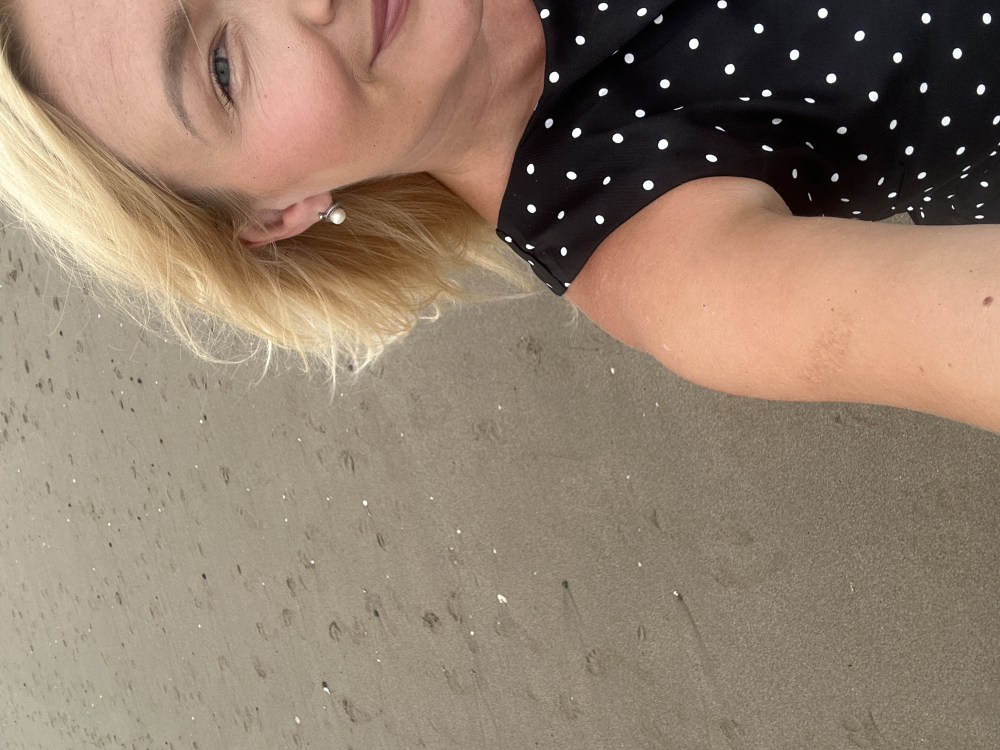 A blonde-haired woman, wearing a black and white polka dot dress and a pearl earring, stands on a sandy beach. The sand is wet and marked with numerous small footprints. The woman is smiling and looking towards the camera. Her hair is blowing gently in the wind. The background is a sandy beach with a muted grey tone, suggesting it is likely overcast or dusk. The location is identified as North Kuta, Indonesia, which implies a coastal environment.